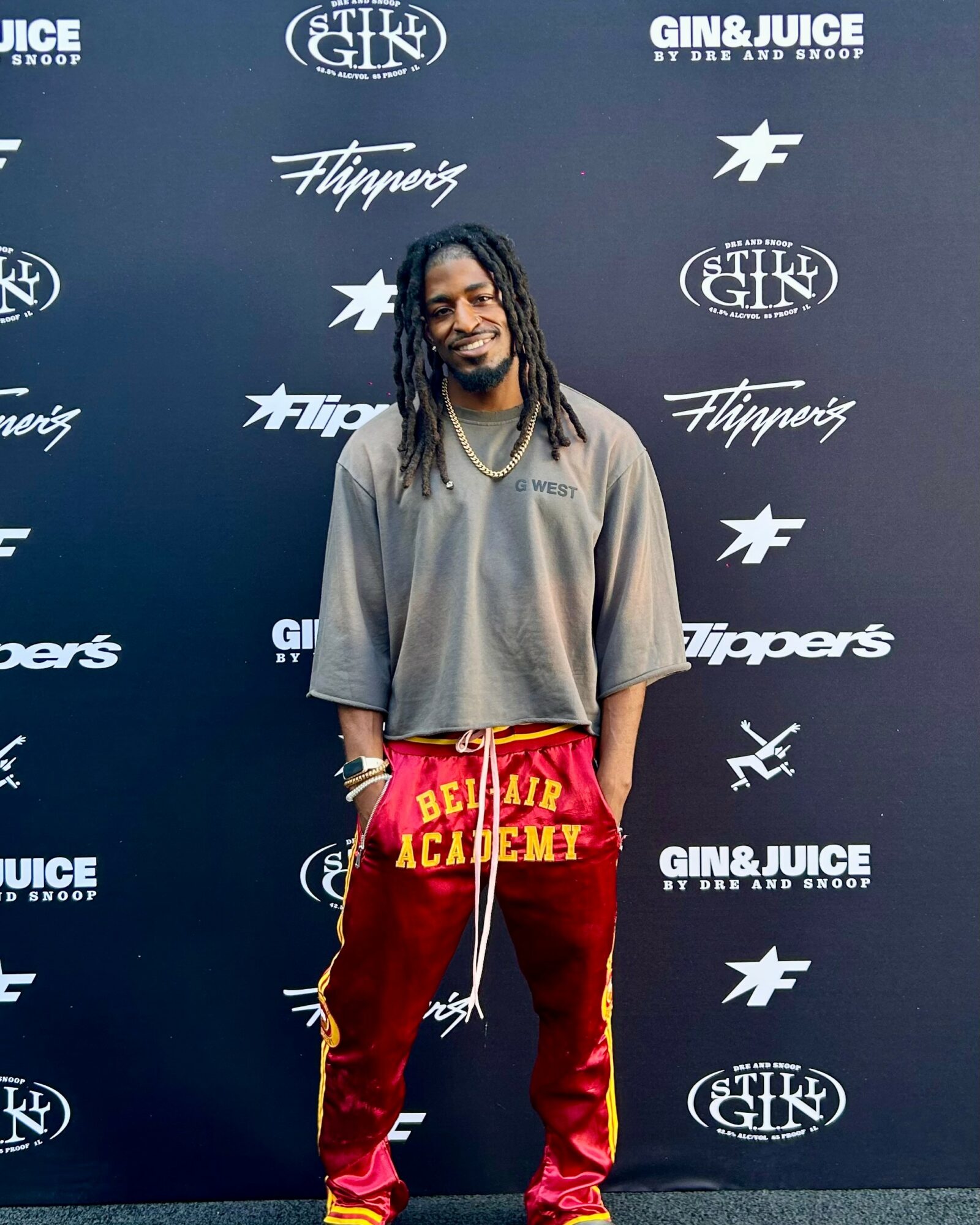 Man with dreadlocks smiling, wearing a gray oversized shirt and red pants, standing in front of a black backdrop with logos.