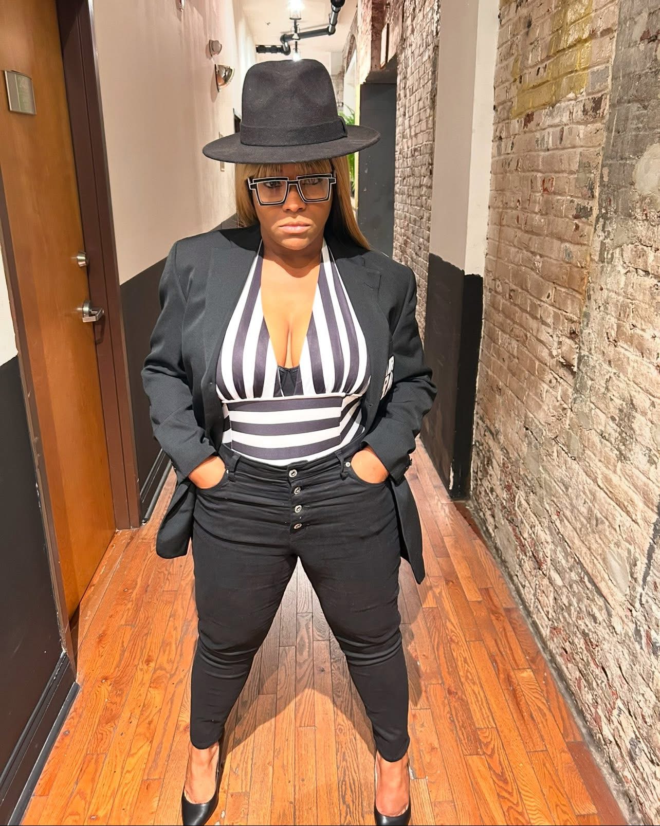 Woman in black blazer, striped top, black pants, black hat, standing in hallway with brick wall, wooden floor.