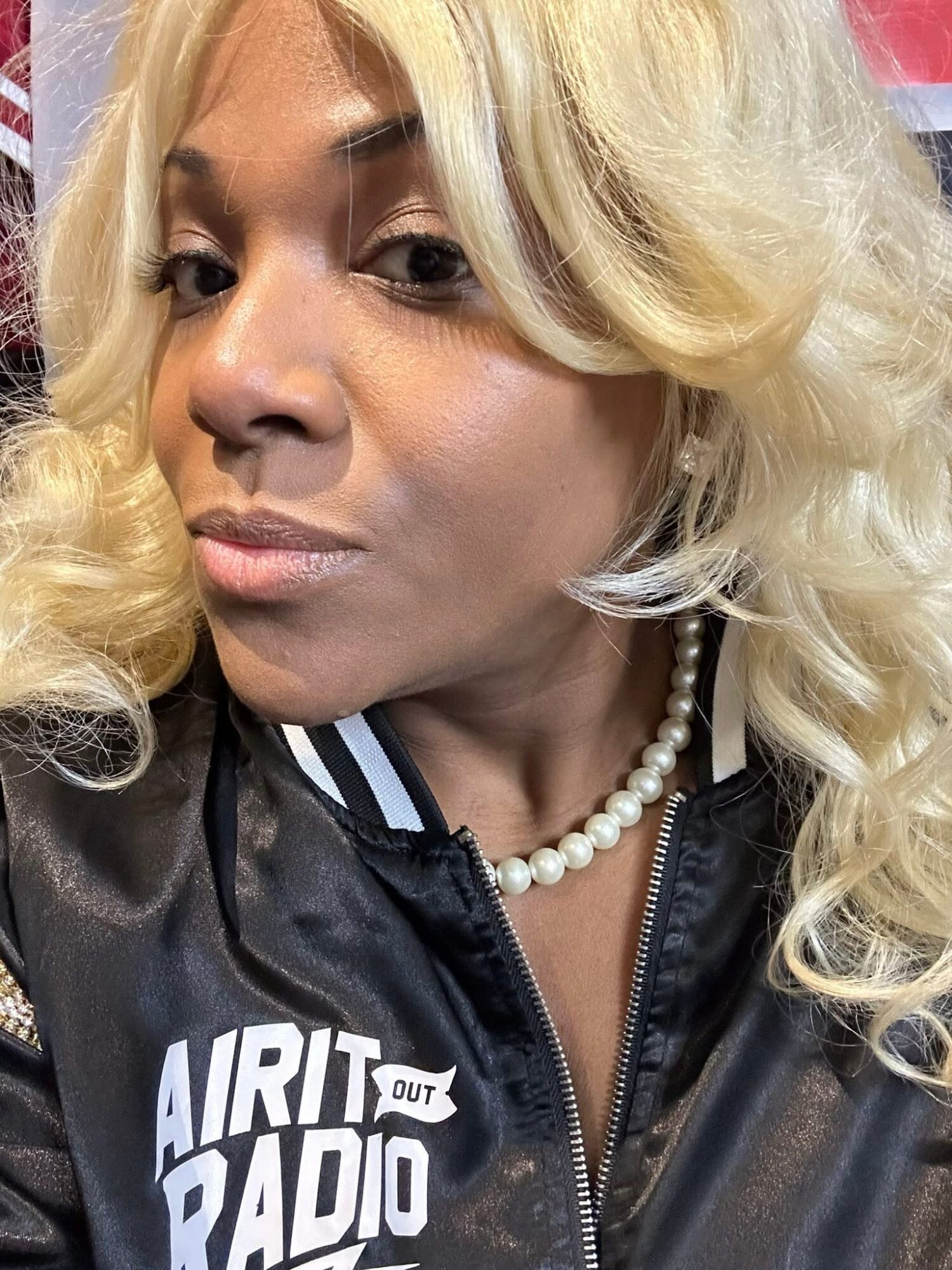 Close-up of a woman with blonde curly hair wearing a black jacket with white text and a pearl necklace.