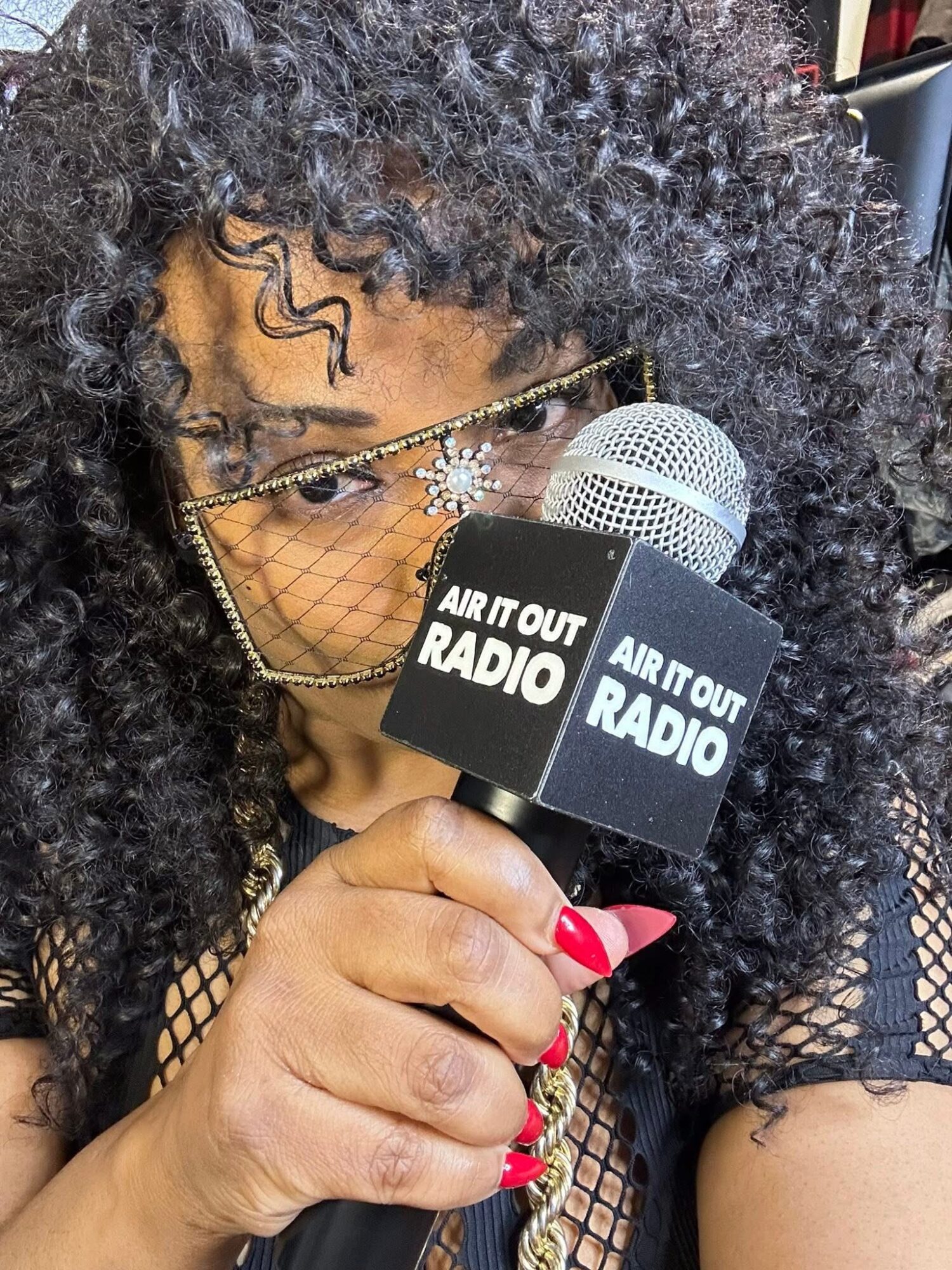 Person with curly hair holding a microphone with 'AIR IT OUT RADIO' logo, wearing glasses and a face mask, close-up.