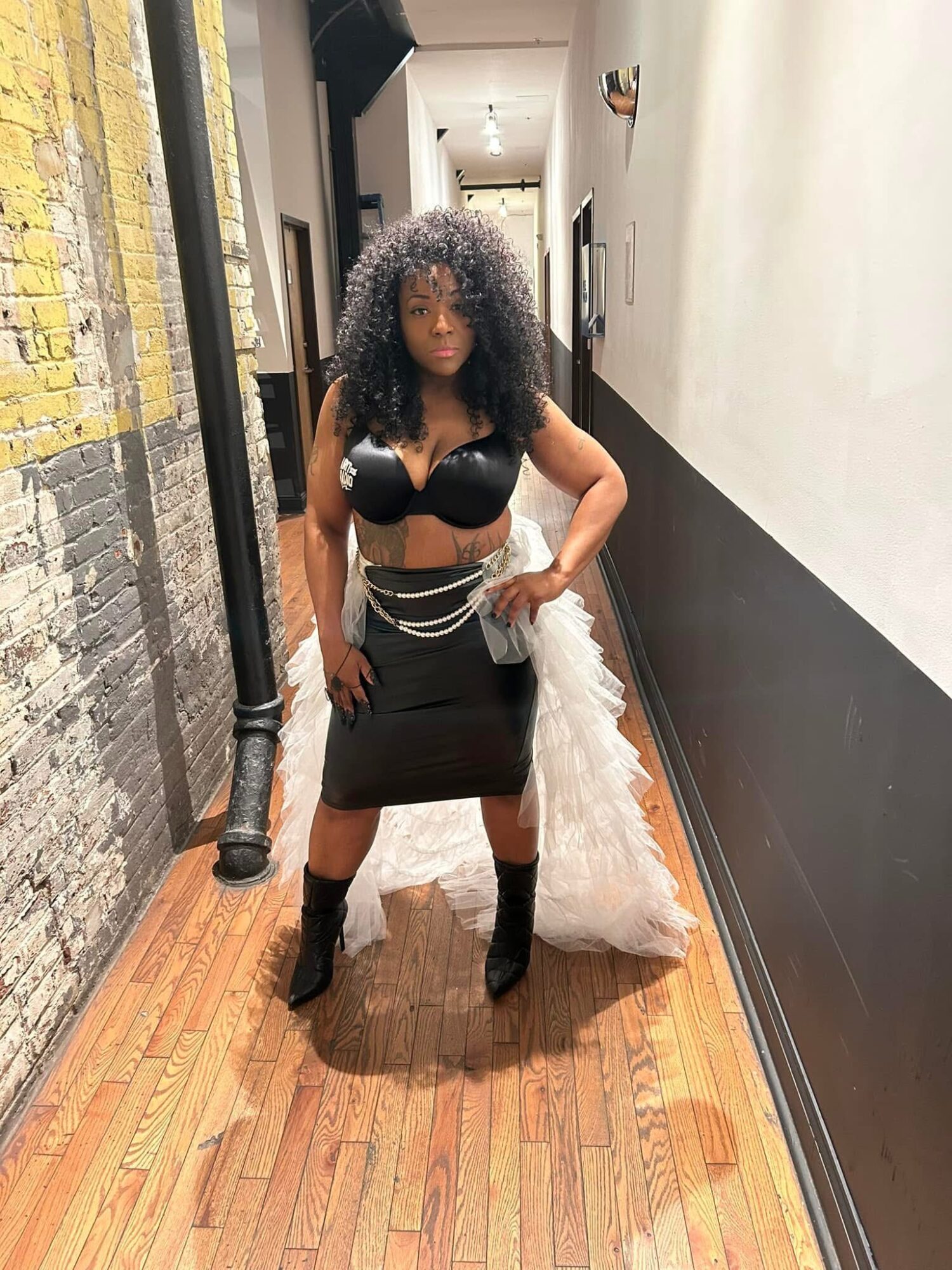 Woman with curly hair in a black outfit and high heels standing in a hallway with brick and painted walls.