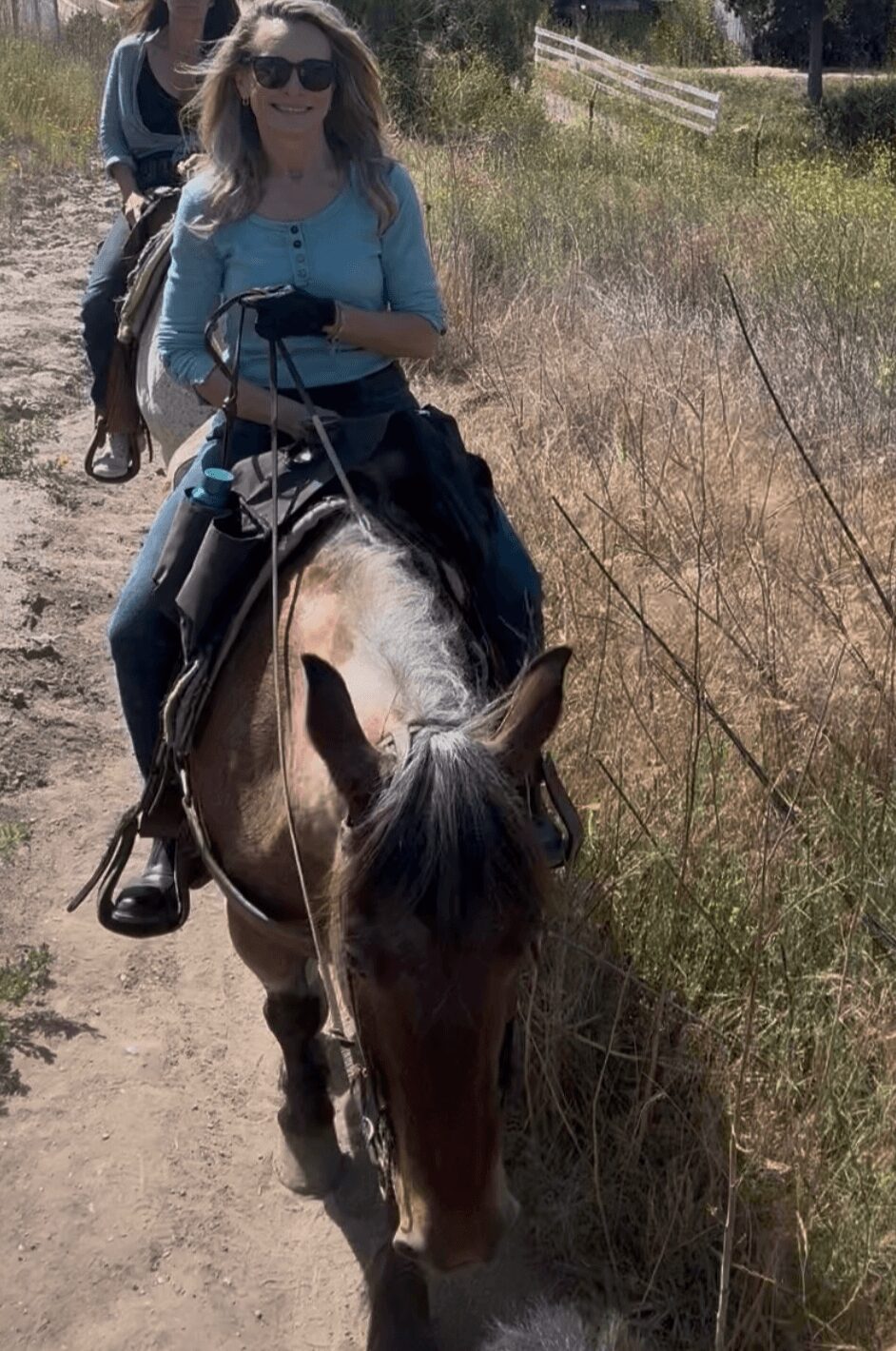 Person riding a horse on a dirt path with grassy surroundings and a wooden fence in the background.