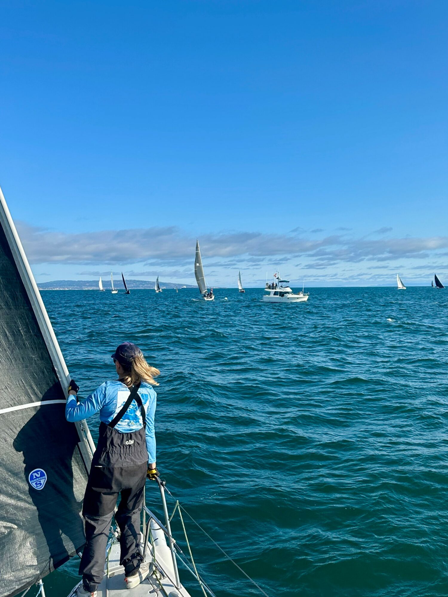 Person standing on a sailboat, holding a rope, with sail and water in the background, under a clear blue sky.