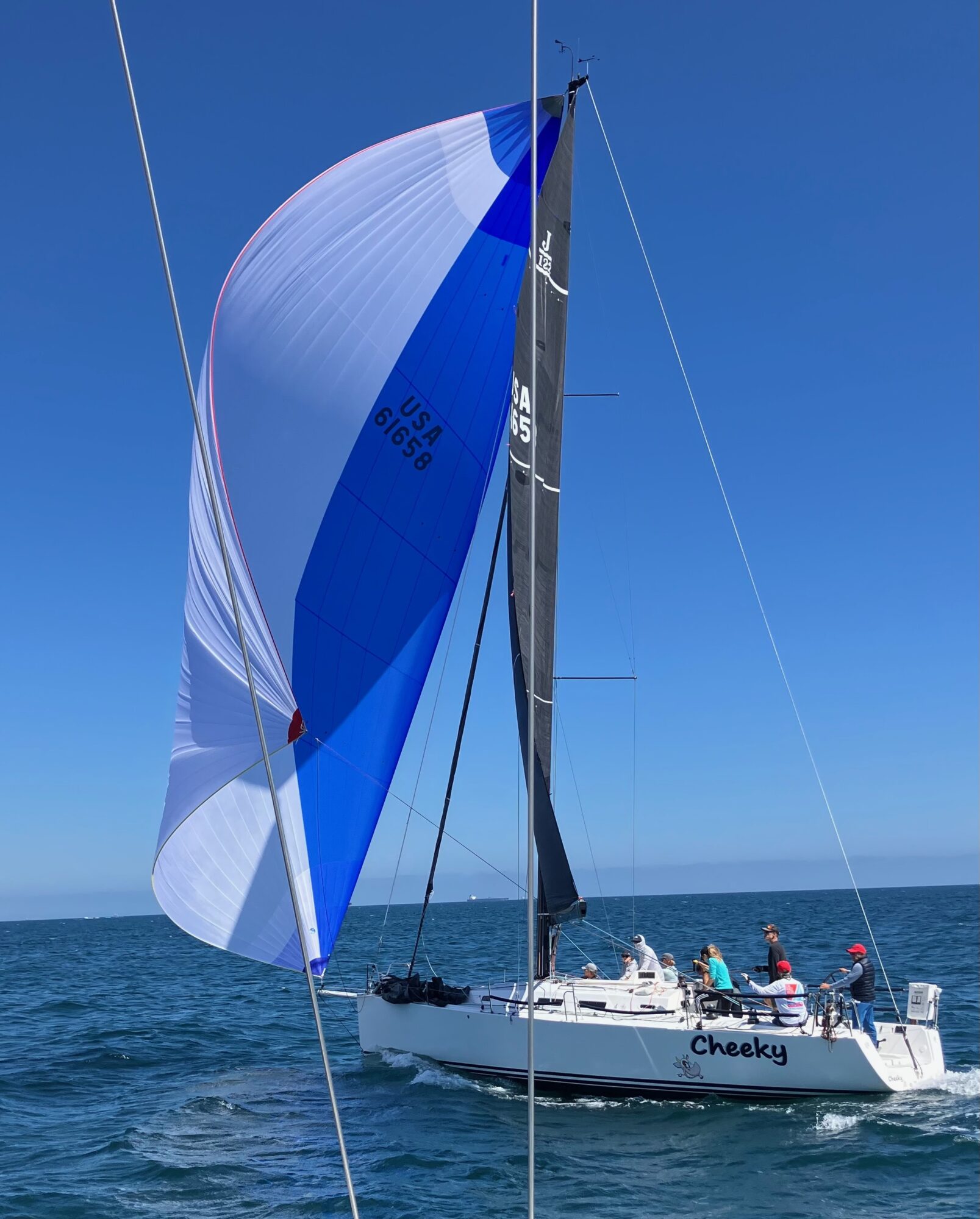 Sailboat with blue and white spinnaker sail on the ocean, crew members on deck, clear blue sky, named Cheeky