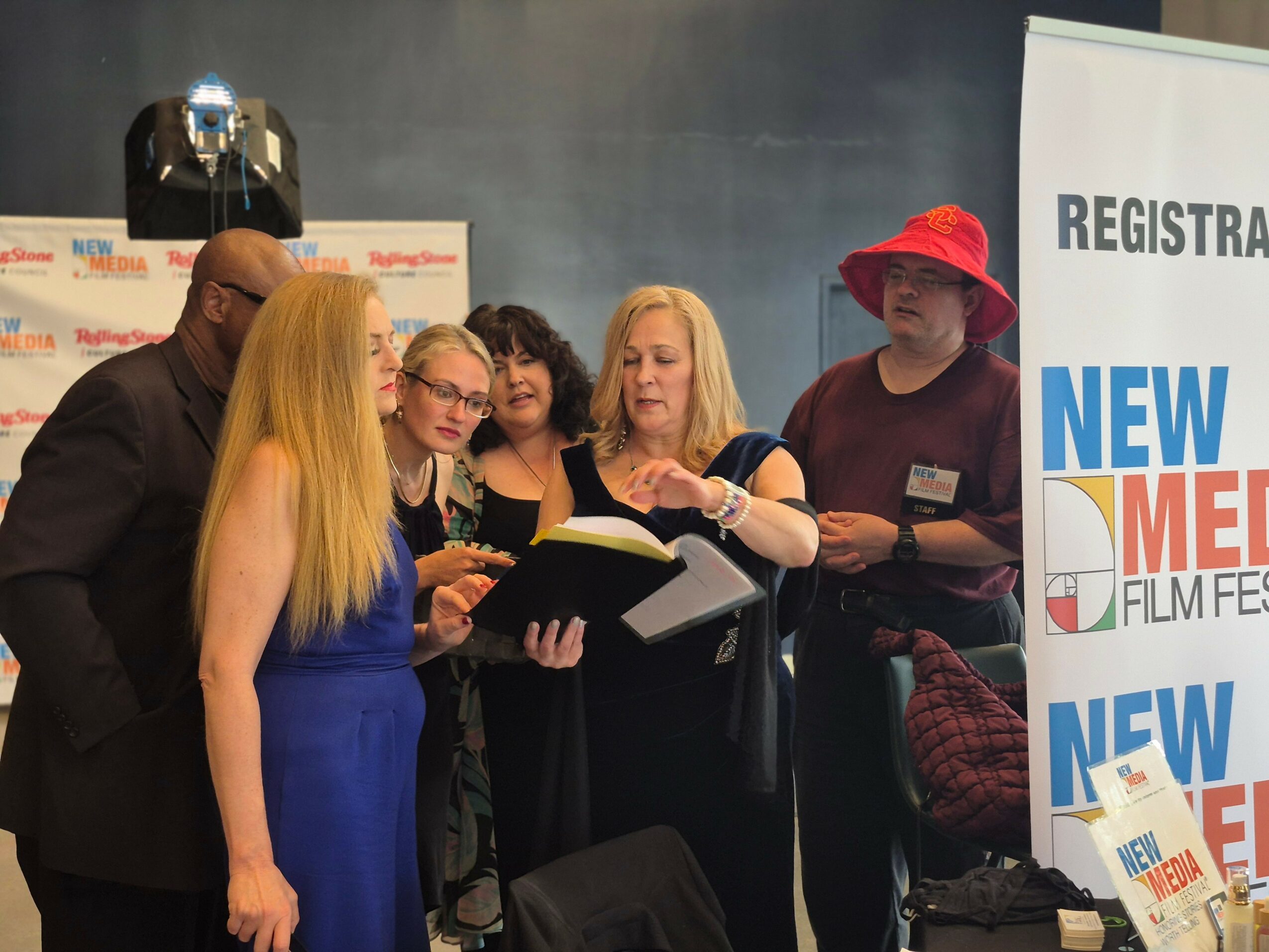 Group of people gathered around a woman reading from a book at an event, with a registration sign in the background.