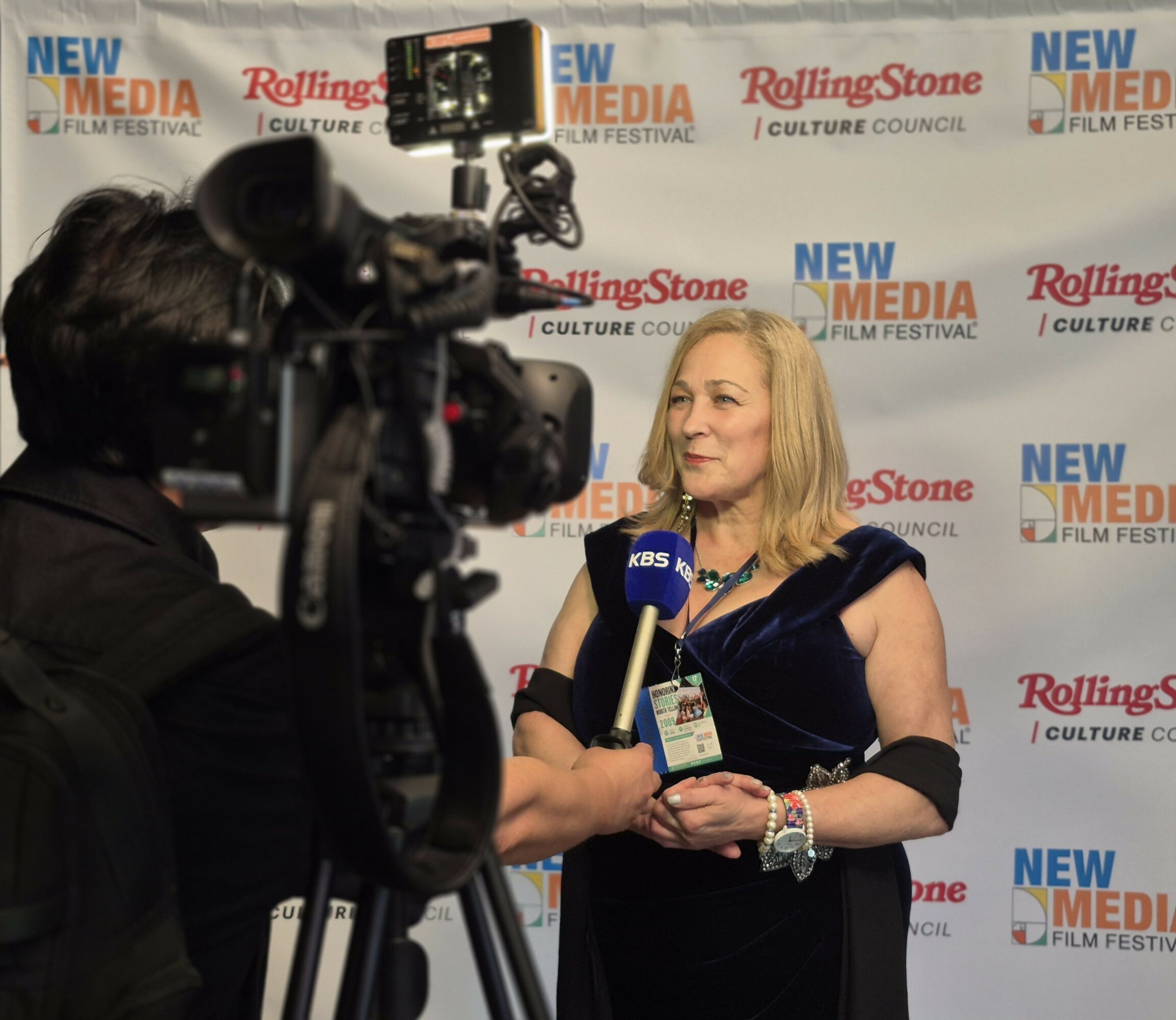 Woman being interviewed on red carpet at New Media Film Festival, holding a microphone, with camera operator filming her.