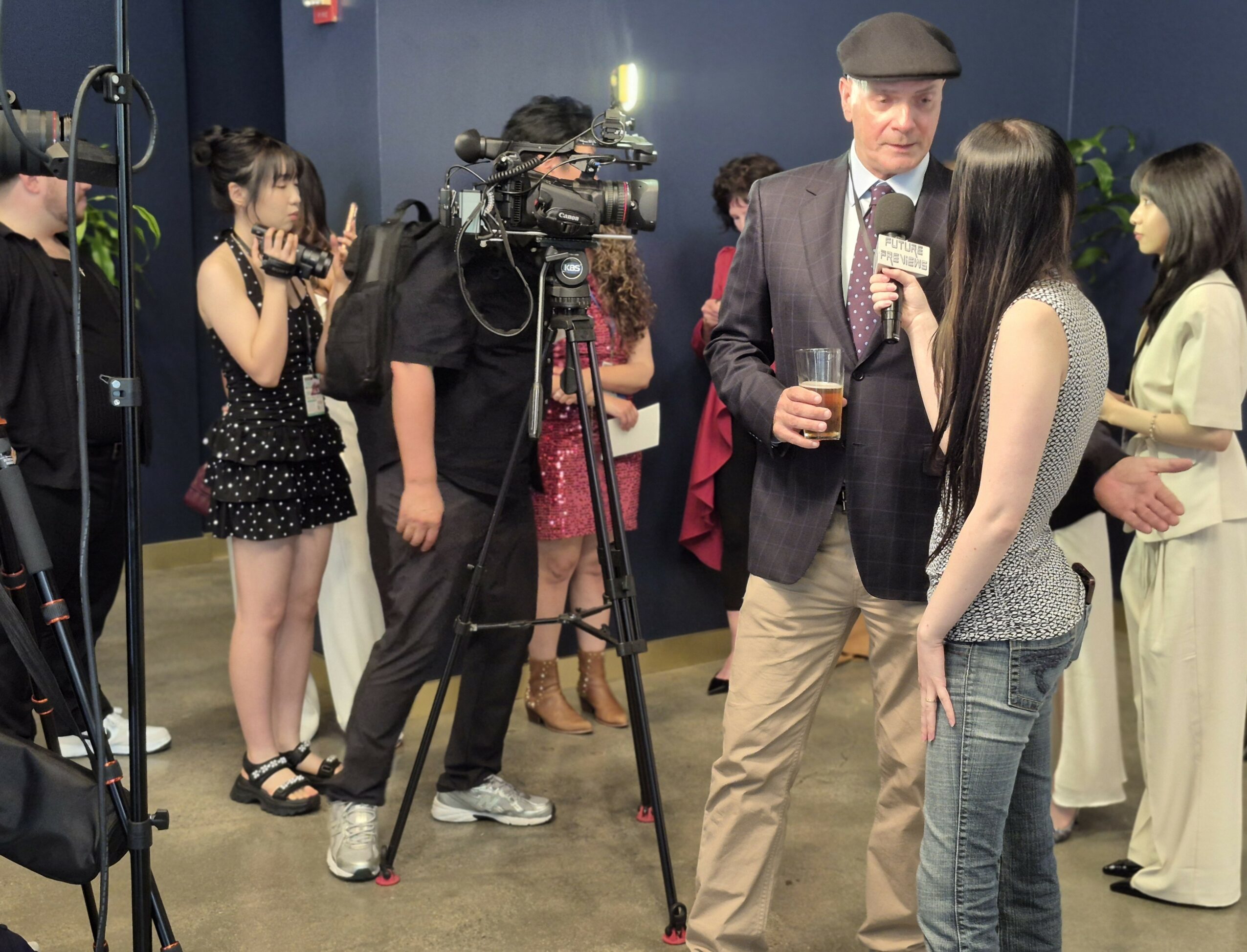 Man in blazer and hat being interviewed by woman with long dark hair, surrounded by people and camera crew in an indoor setting.