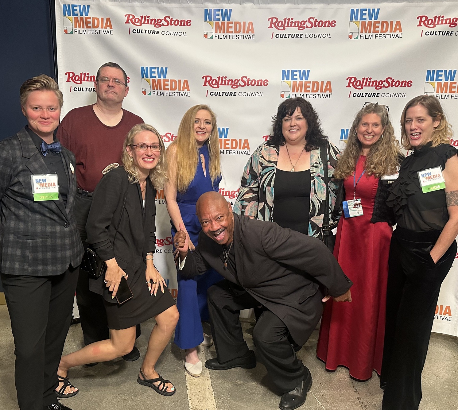 Group of nine people smiling and posing in front of a backdrop with event logos, some wearing name tags.