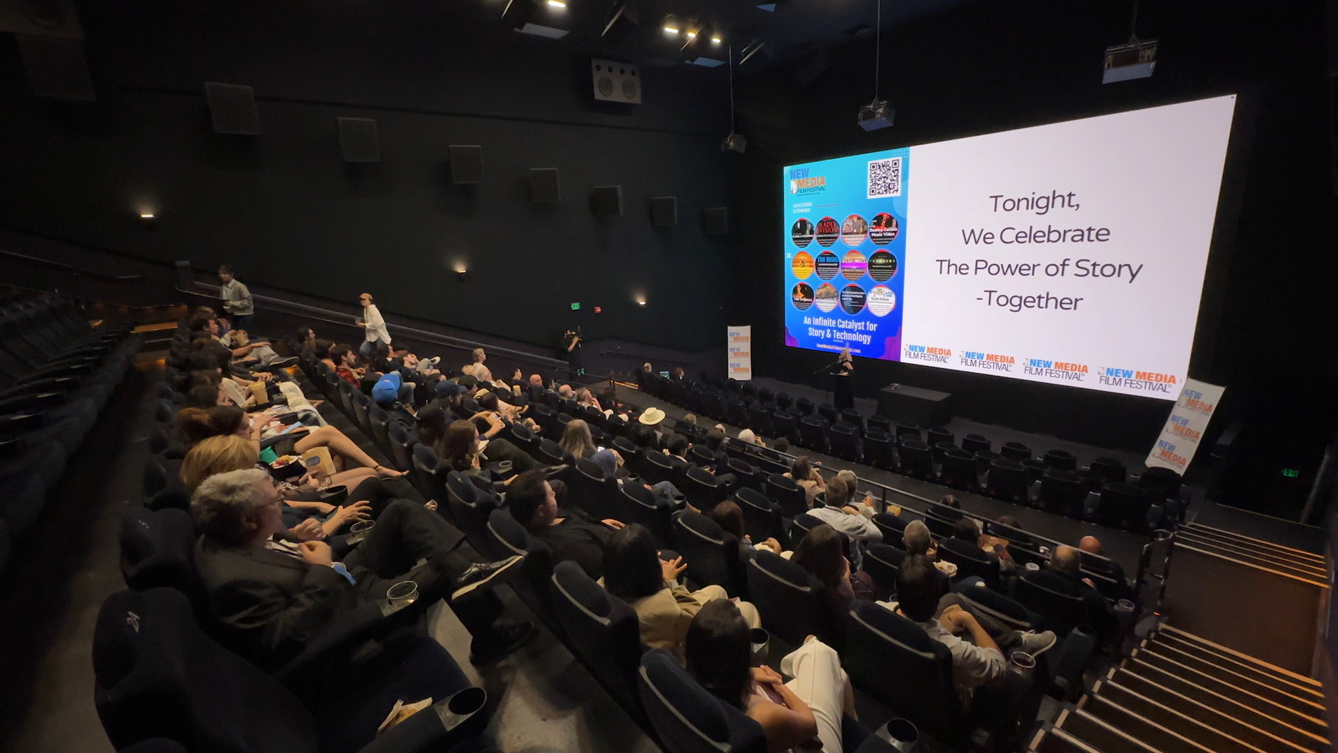 Audience seated in a theater watching a presentation on a large screen, with some people taking photos.