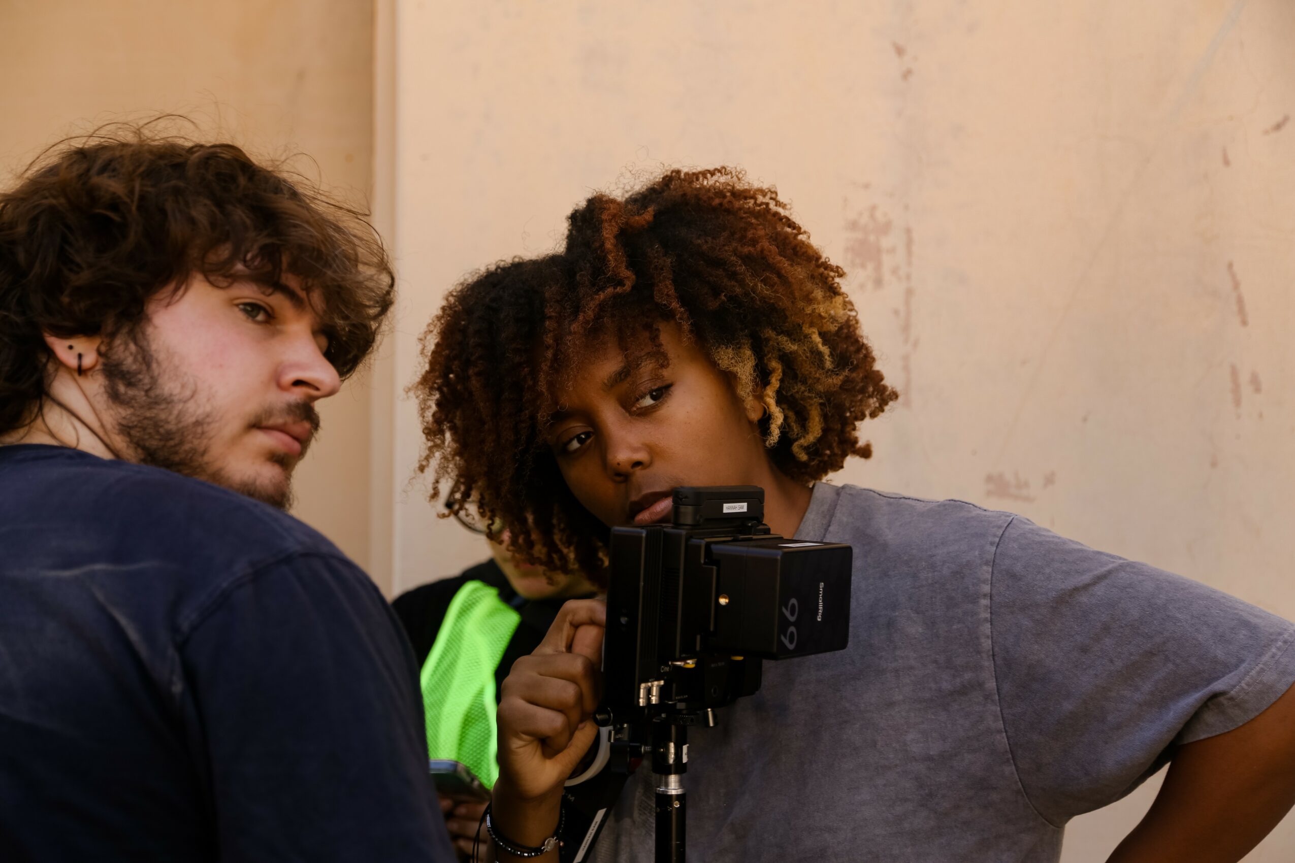 Two people looking at a camera on a tripod, one with curly hair and the other with dreadlocks, in a room.
