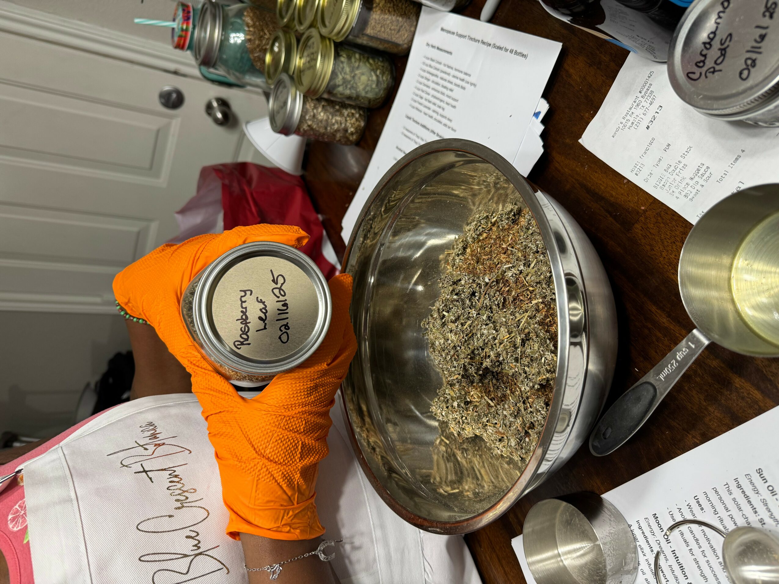 Person wearing orange glove holding a small container over a metal bowl filled with a mixture of herbs and spices, on a cluttered table.