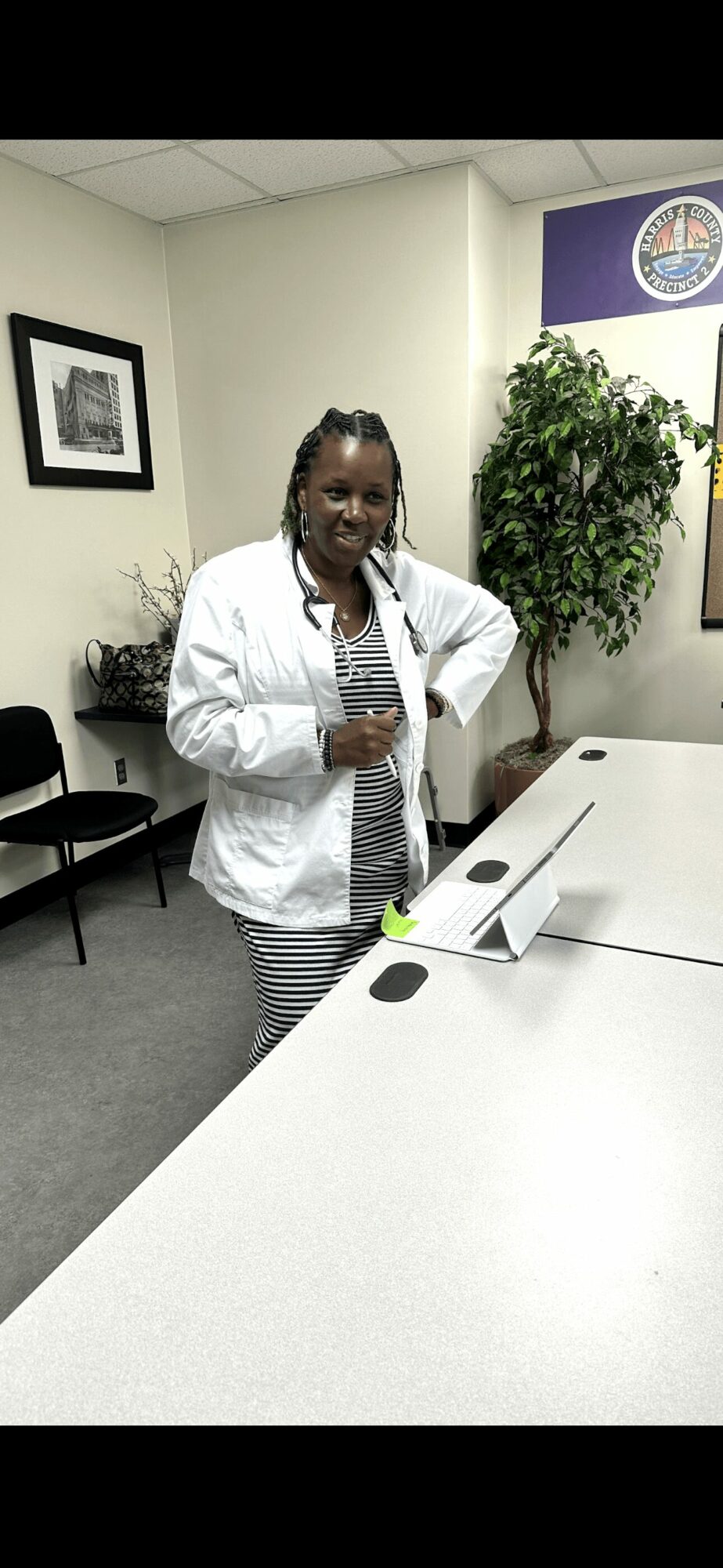 Woman standing at a table in an office, smiling, with a plant and framed pictures on the wall behind her.