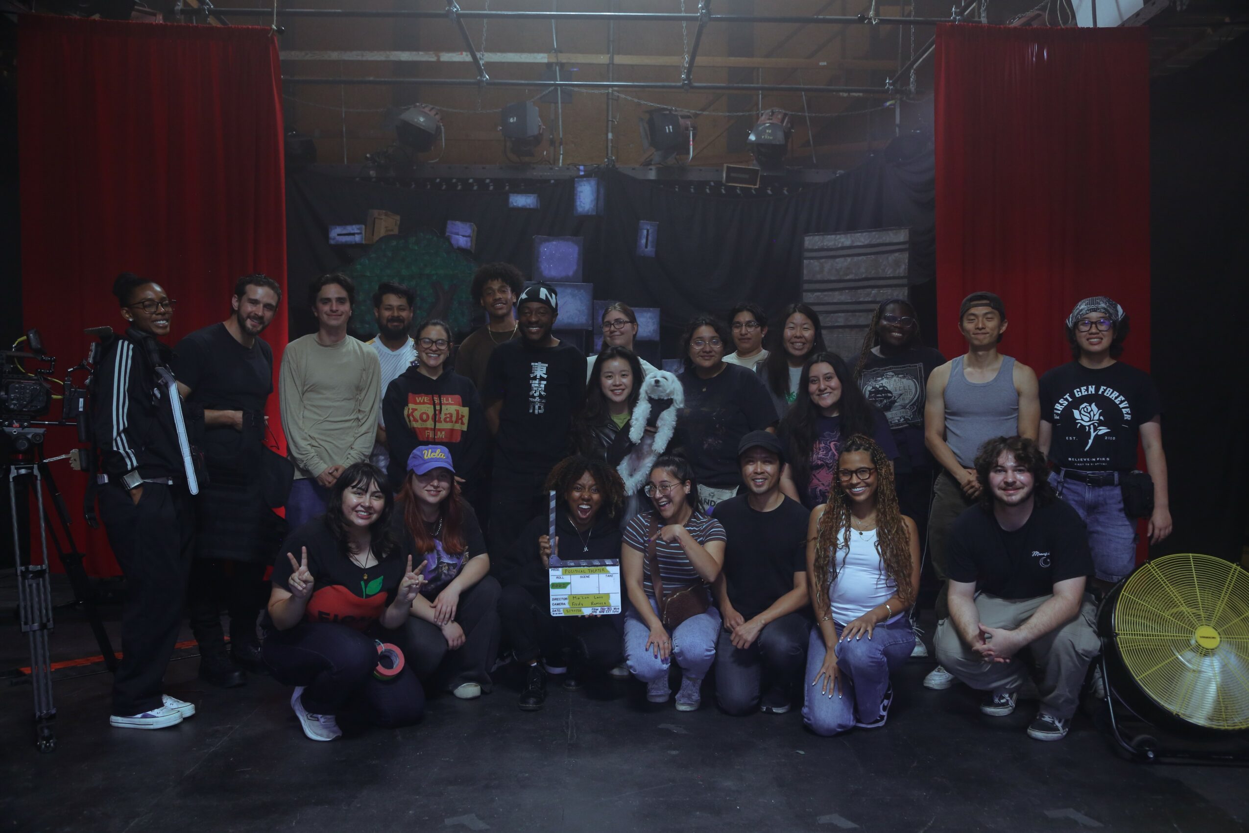 Group of people posing on stage with red curtains and equipment in background, some holding a certificate or paper.
