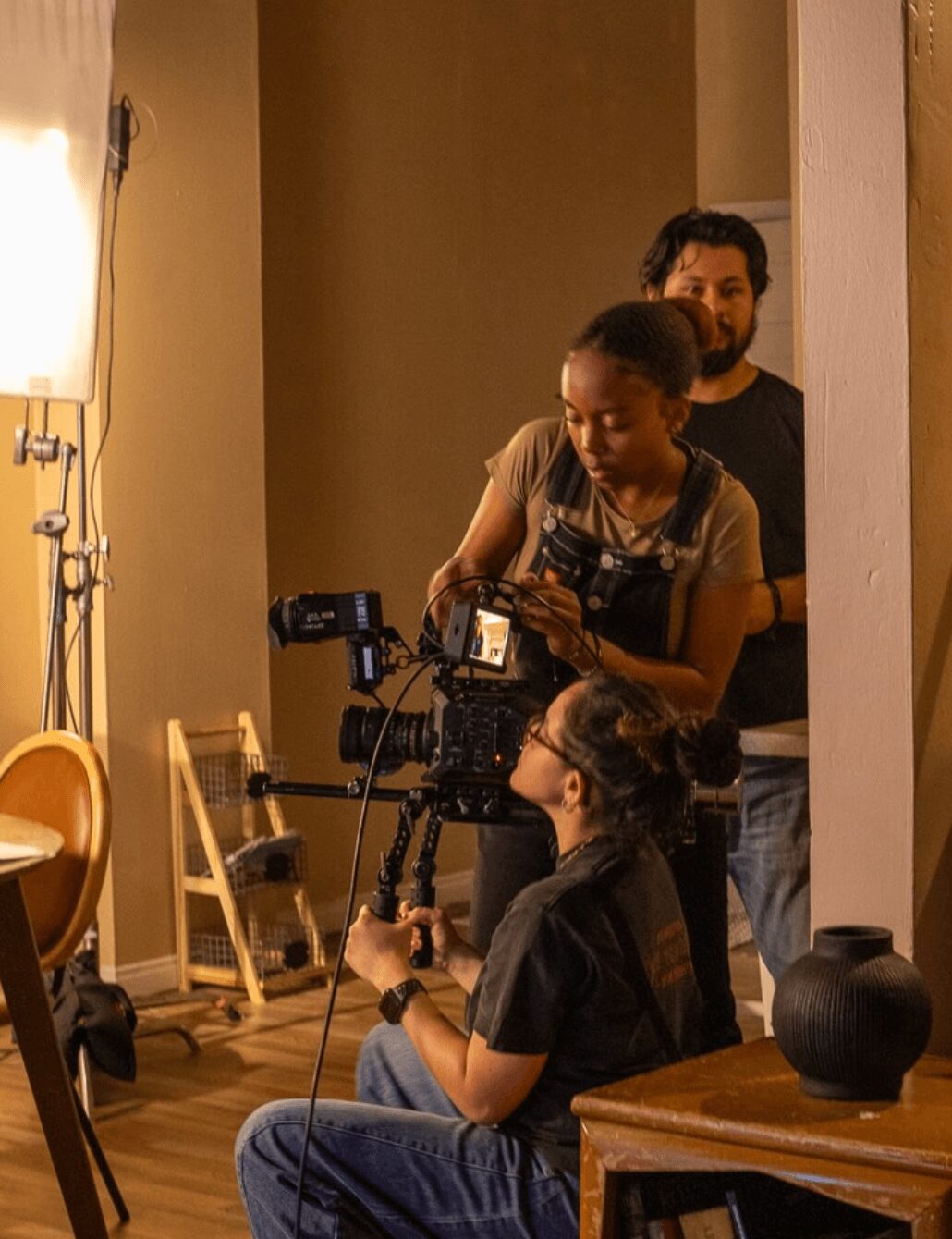 Three people working with camera equipment indoors, one person sitting and two standing, in a room with warm lighting.