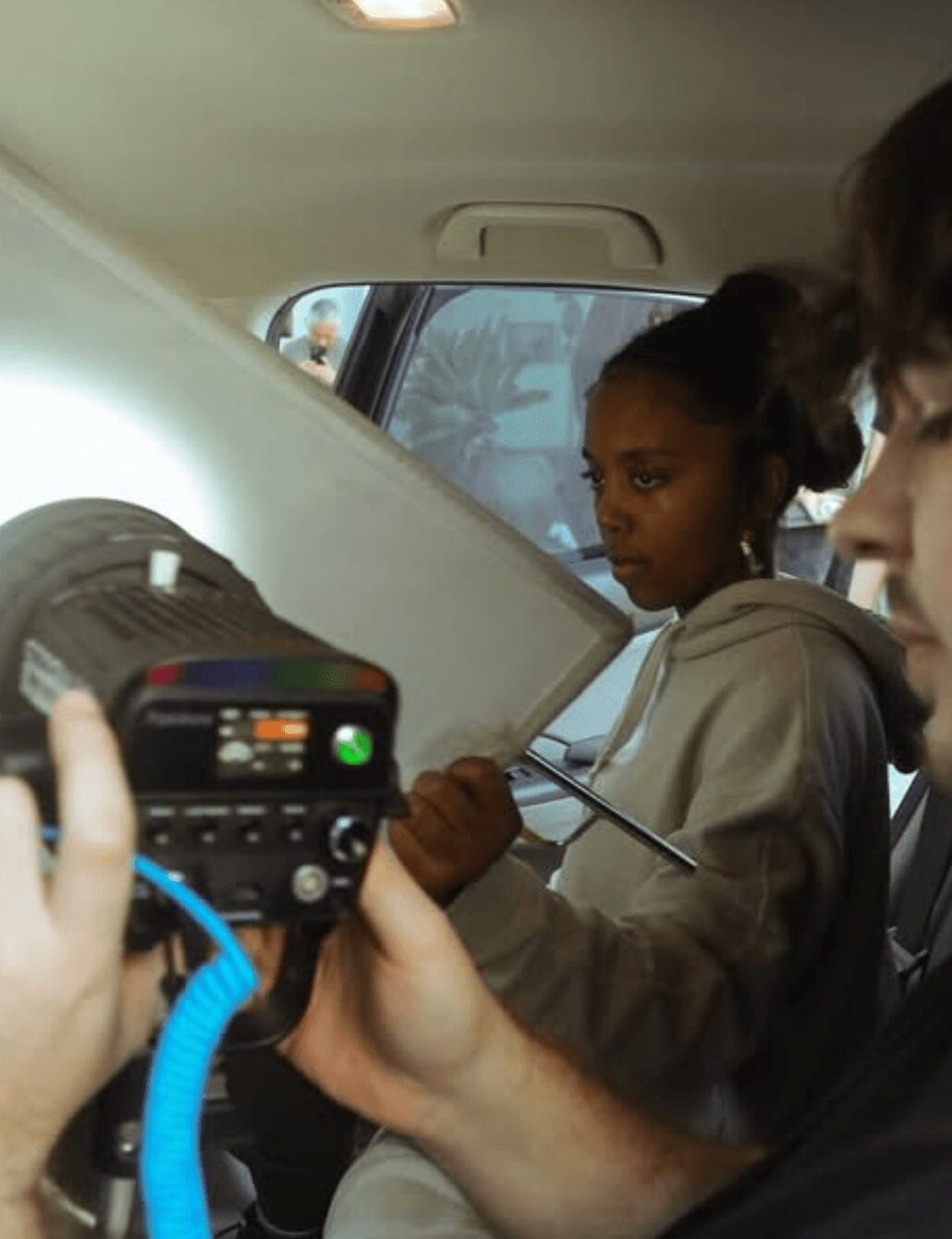 Two people inside a vehicle, one holding a camera, and a girl looking at a large white object, possibly a board or screen.