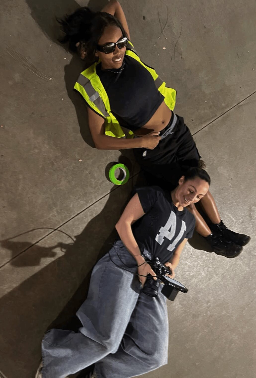 Two women lying on the floor, one with sunglasses and a yellow vest, the other holding a camera, both smiling.
