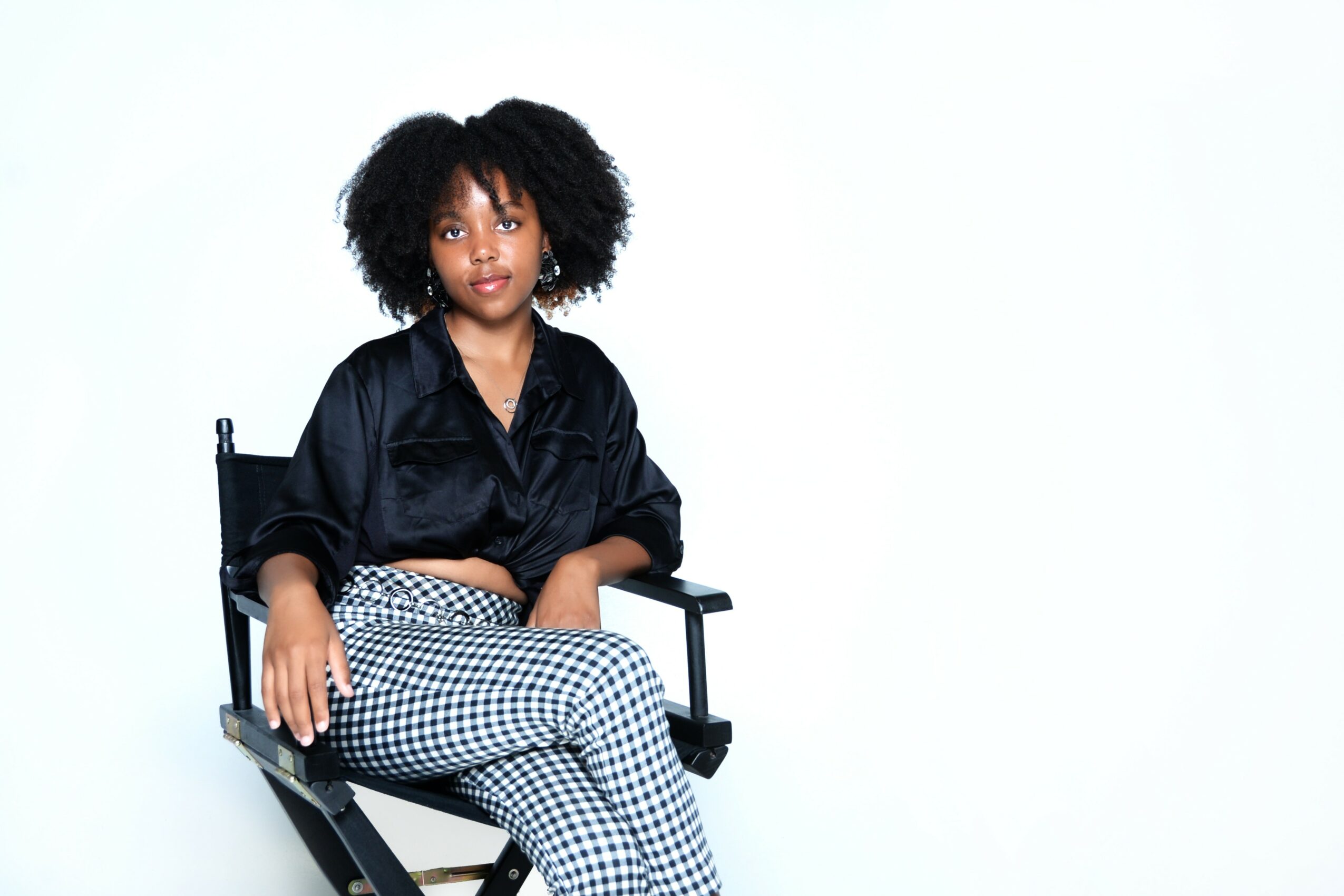 Young woman with curly hair sitting on a chair, wearing a black top and checkered pants, against a white background.