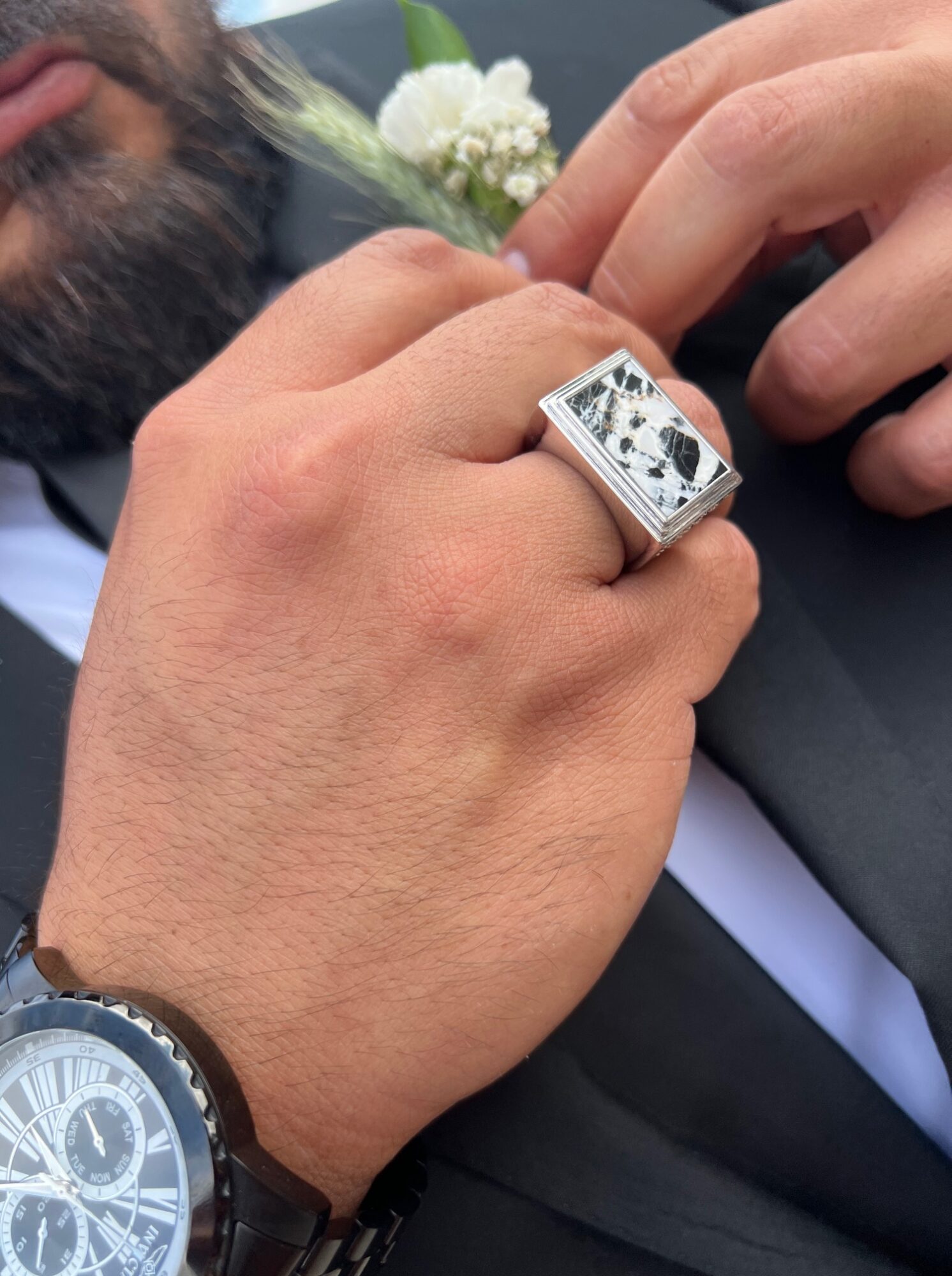 Close-up of a person's hand wearing a large silver ring and a black watch, with a boutonniere in the background.
