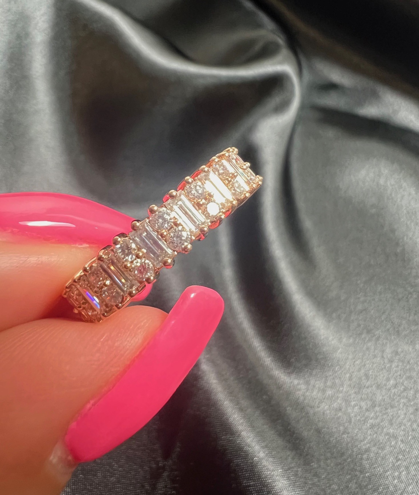 Person holding a diamond ring with a black satin background, pink nail polish, and a shiny surface.