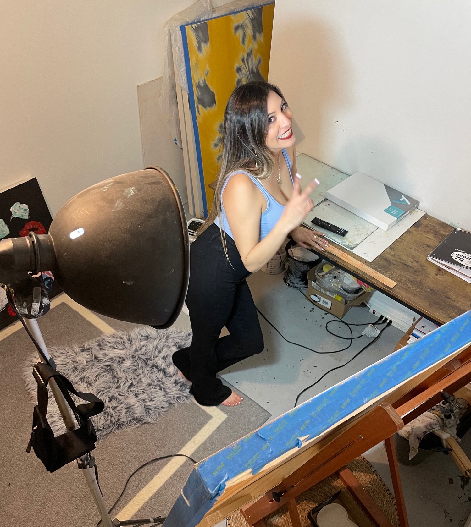 Young woman with long hair in a sleeveless top and dark pants, smiling and making a peace sign, in a room with art supplies.
