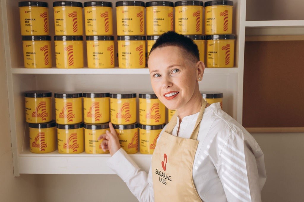 Woman in apron standing in front of shelves filled with yellow containers, pointing at one container, smiling.