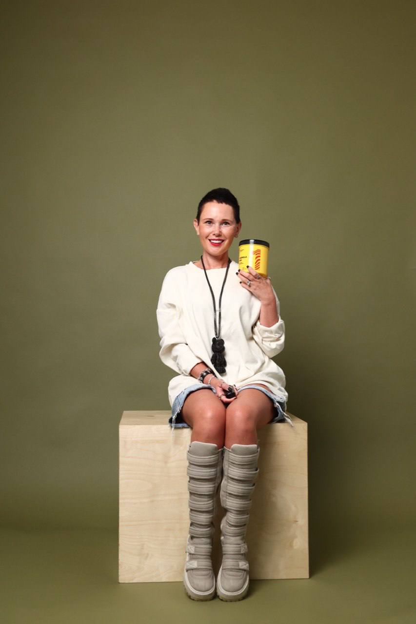 Woman sitting on a wooden block, smiling, holding a yellow cup, wearing a white sweater and striped socks, against a plain green background.