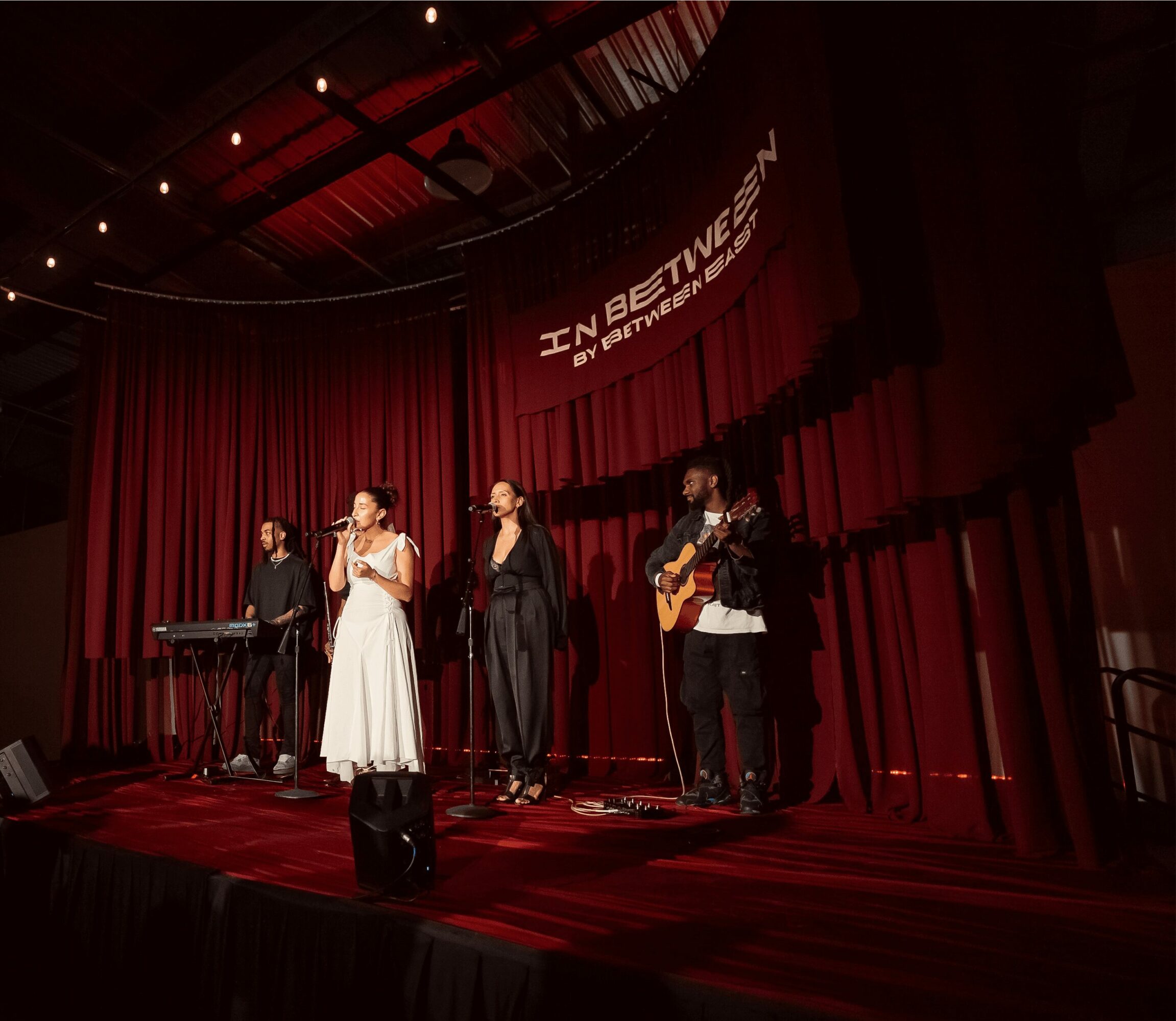 Four performers on stage with red curtains, one playing keyboard, one singing, one with microphone, one playing guitar, stage lit.