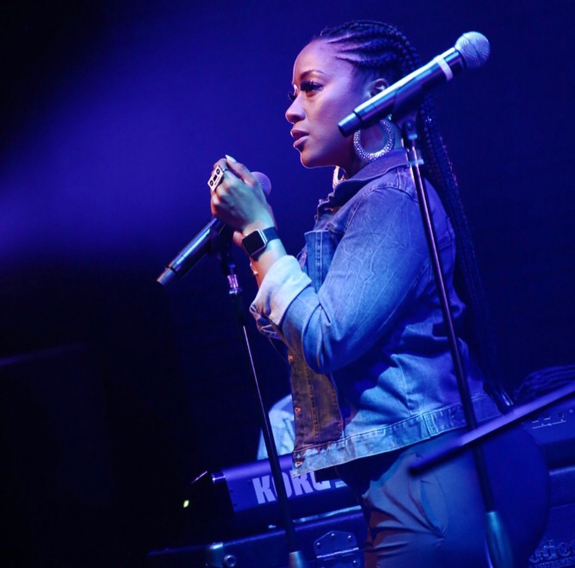 Woman with braided hair holding a microphone on stage, wearing a denim jacket, with a keyboard nearby, under blue lighting.