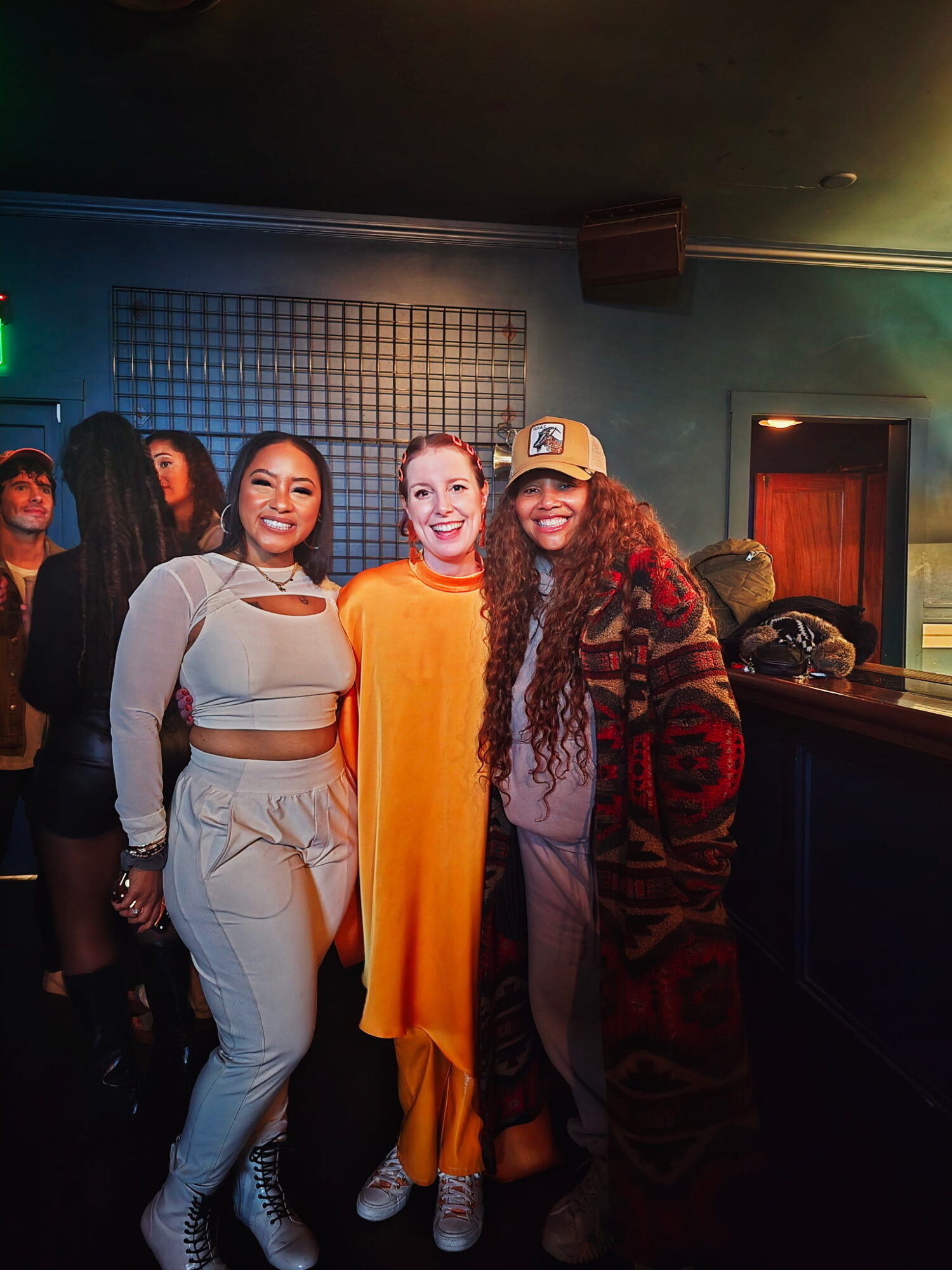 Three women standing together indoors, smiling, with a dark background and other people in the background.