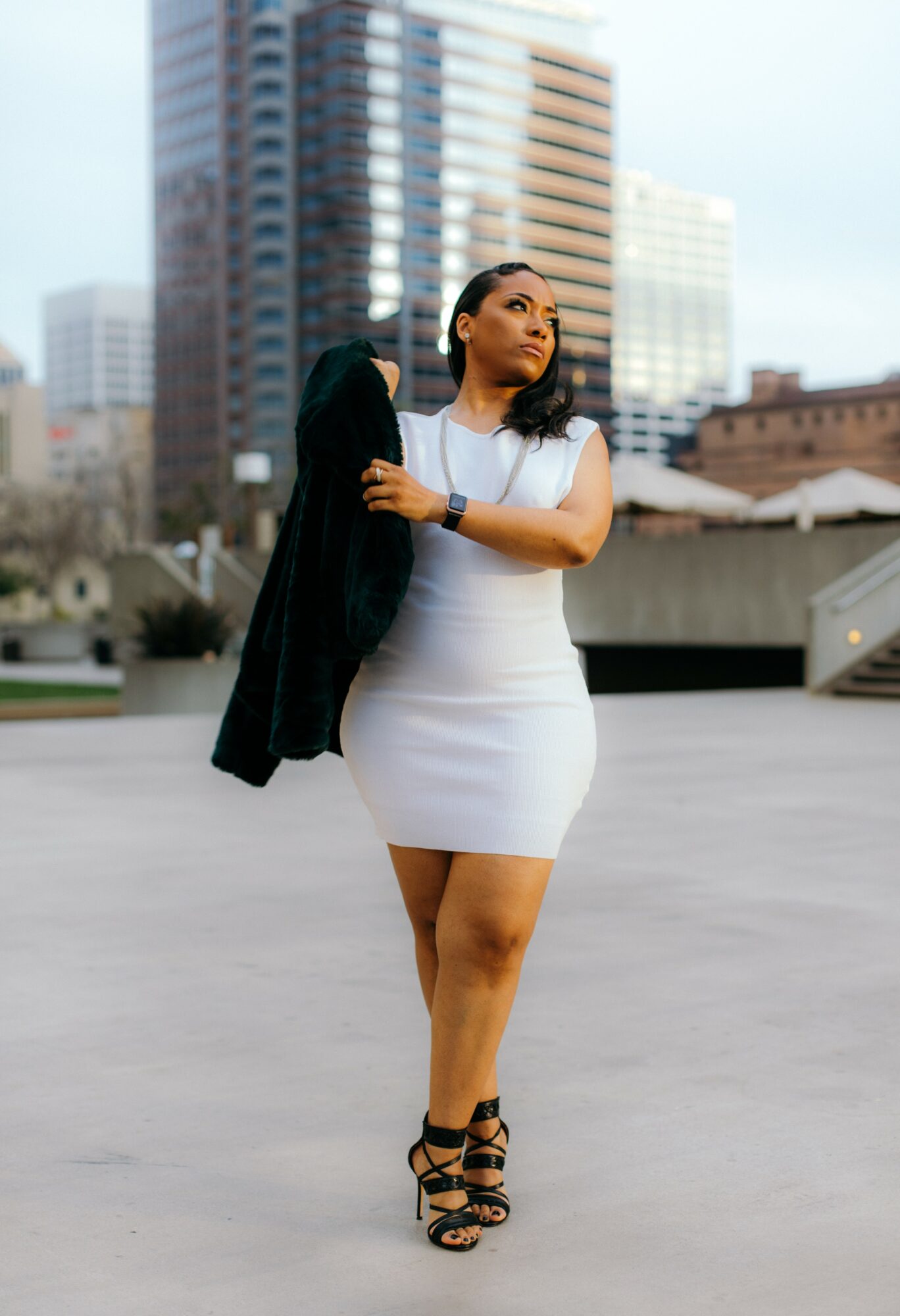 Woman in white dress holding black jacket, standing outdoors in city with tall buildings in background.