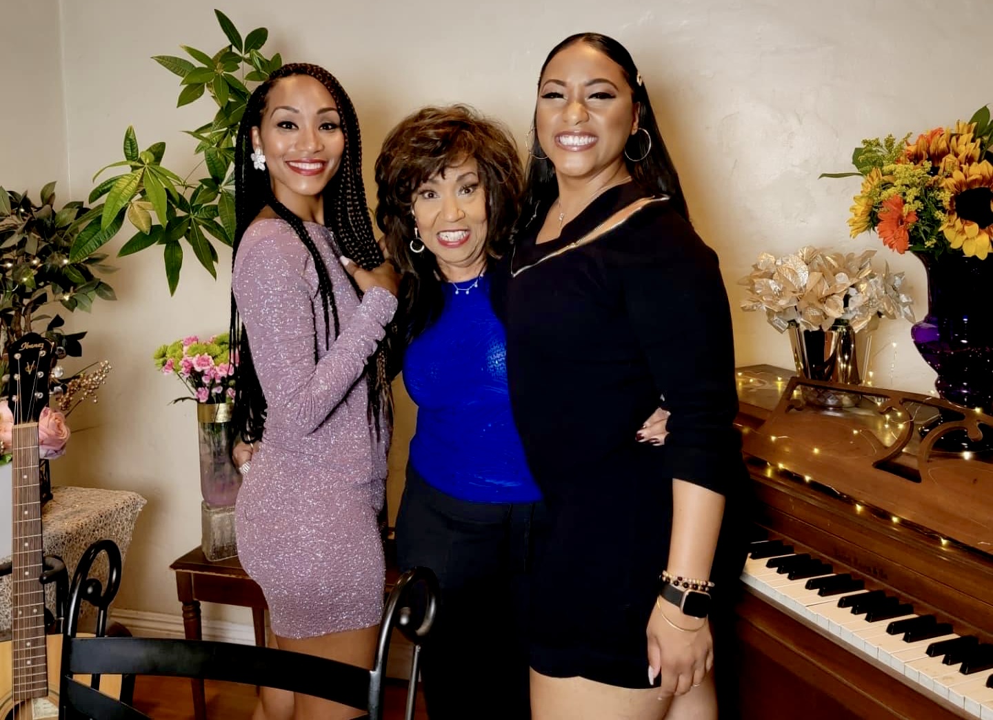 Three women standing together indoors, smiling, with plants and a piano in the background.