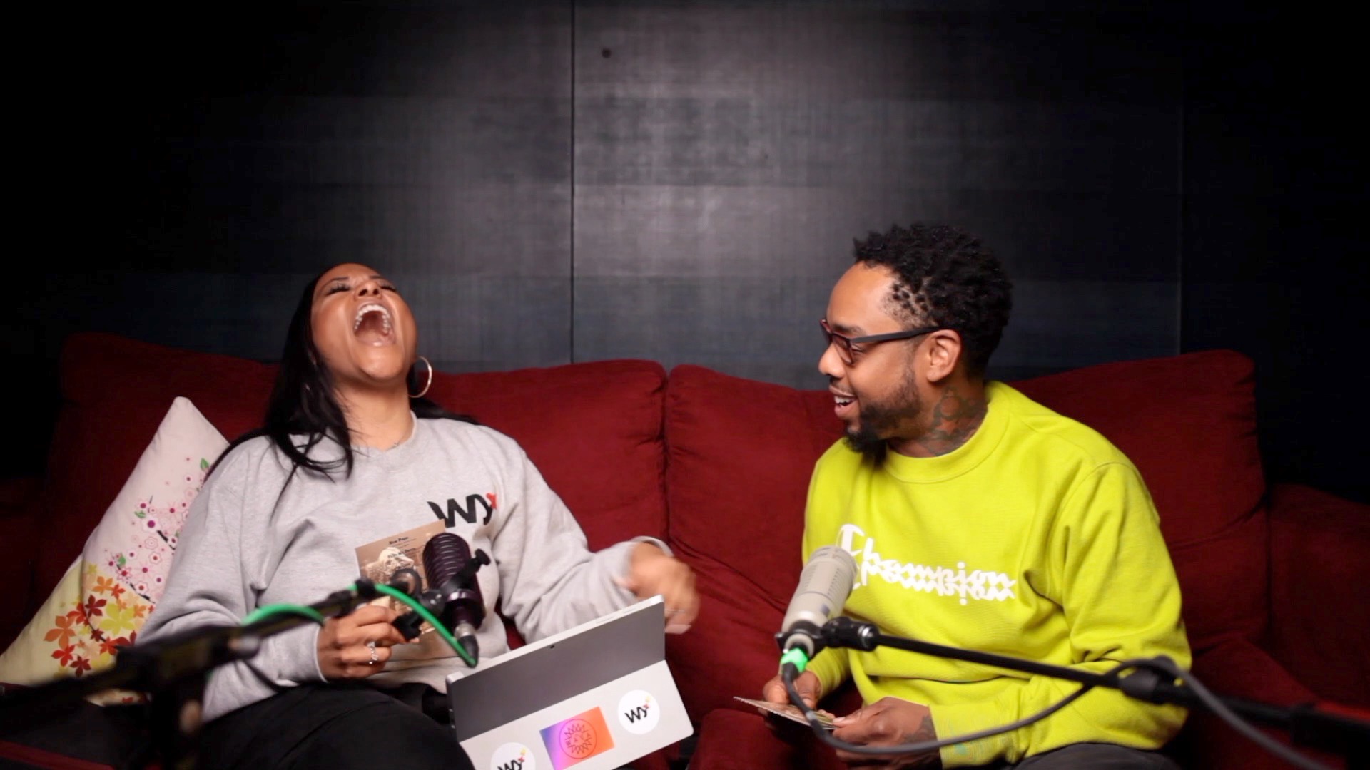 Two people sitting on a red couch, laughing and talking, with microphones and a laptop, against a dark background.