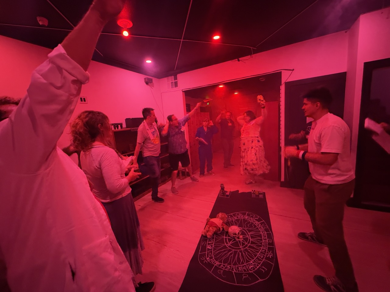 People dancing and socializing in a dimly lit room with red lighting, some raising hands, and a table with a cloth in the foreground.