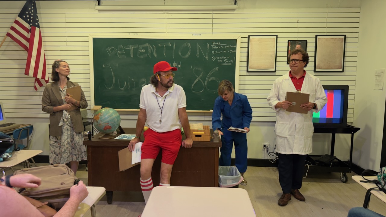 Four people in a classroom with a chalkboard, American flag, and a teacher holding a clipboard, students in costumes, and a TV screen.