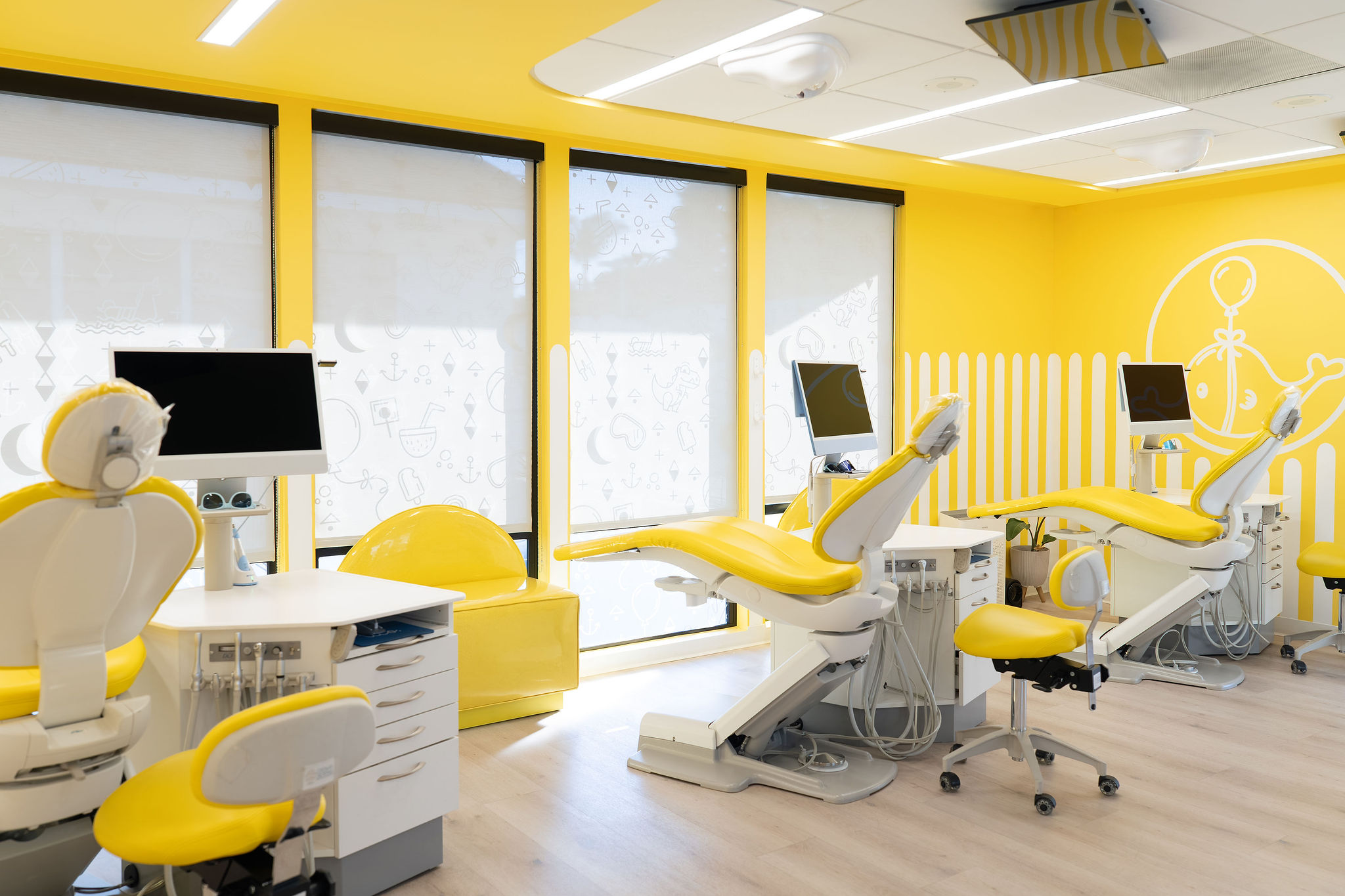 Medical examination room with yellow walls, three examination chairs, and computer monitors, bright natural light from large windows.