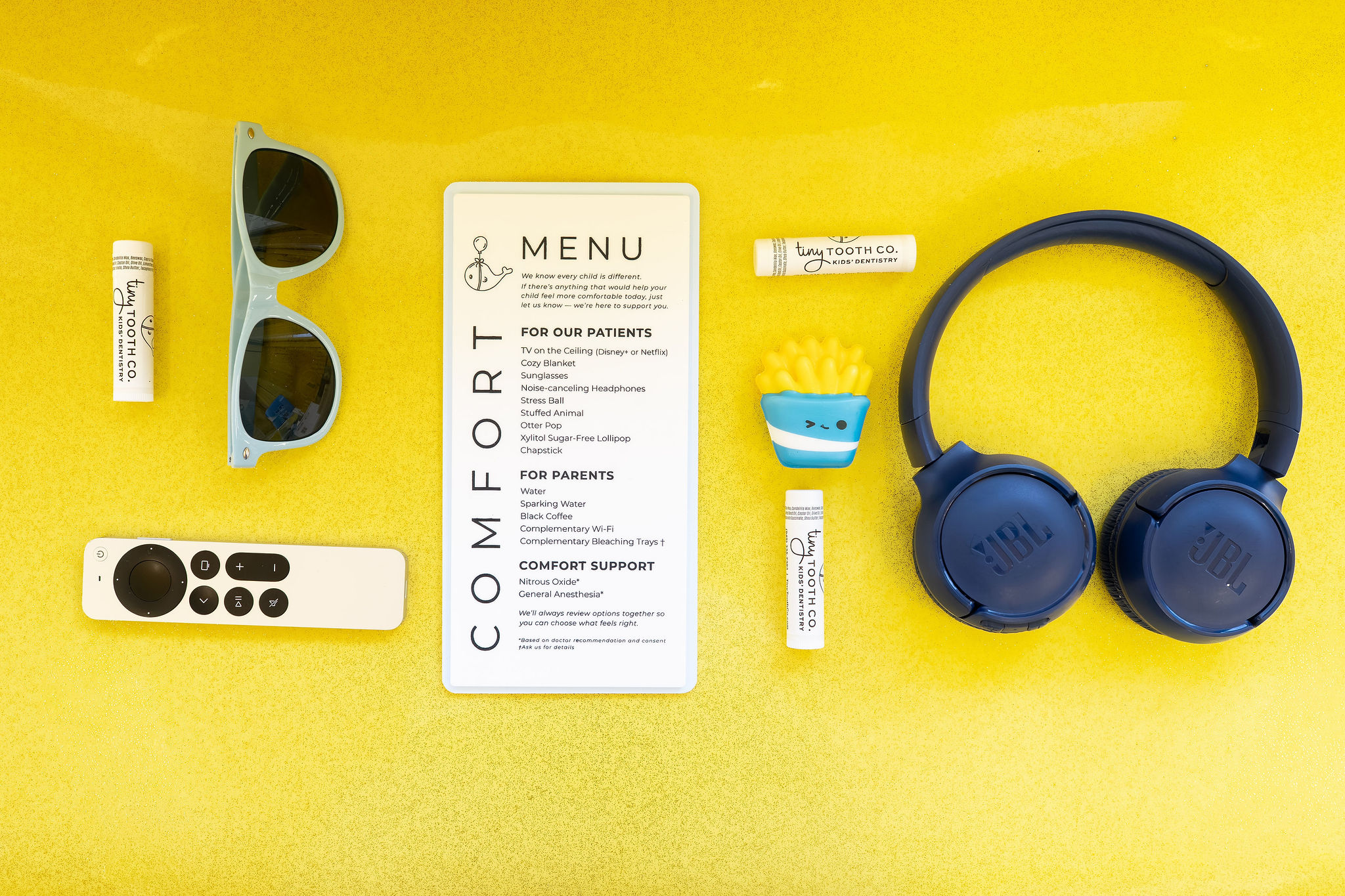 Sunglasses, remote control, menu, earplugs, headphones, and small items on a yellow surface.