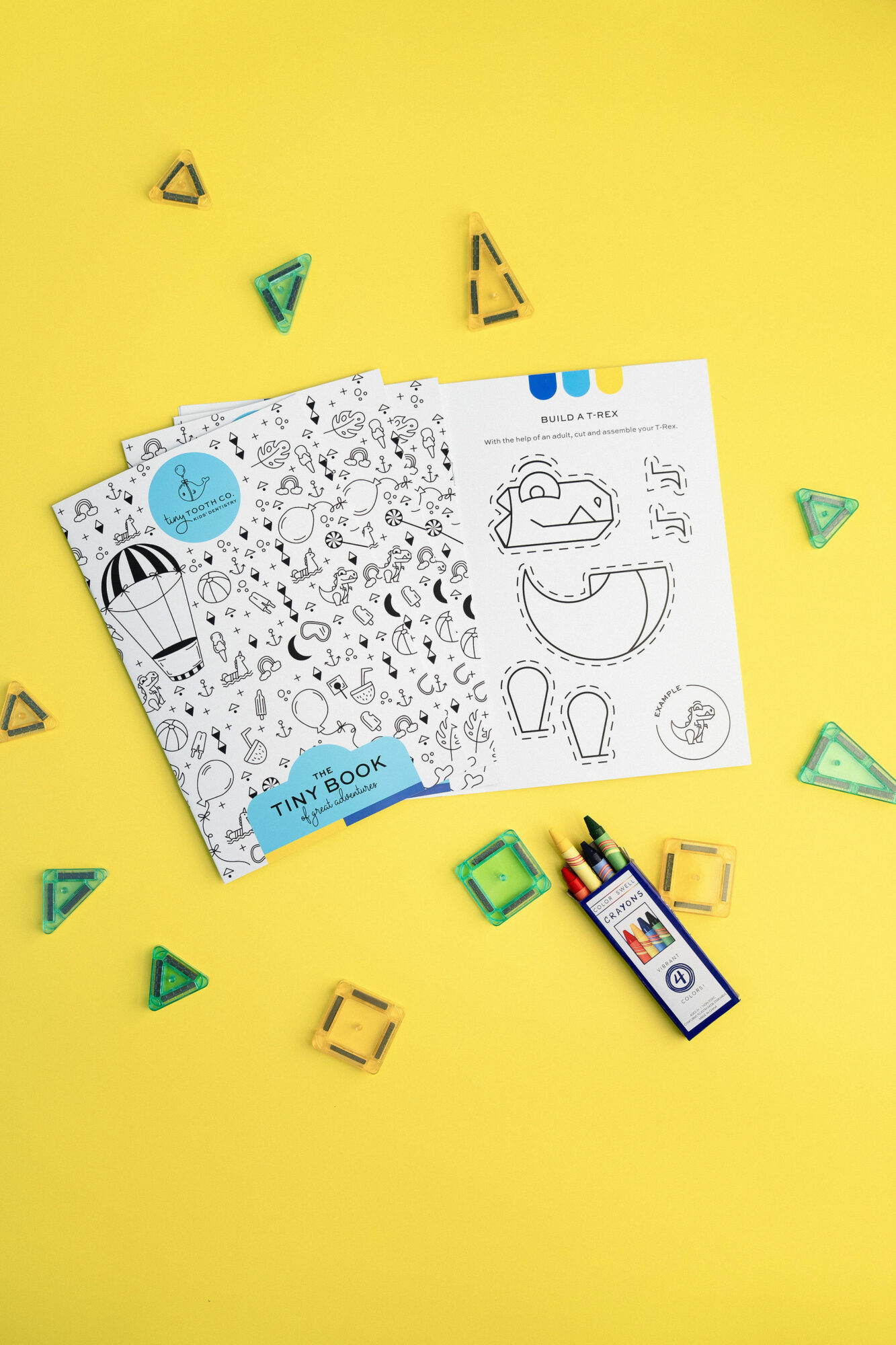 Open activity book with black and white illustrations on a yellow background, surrounded by small green and yellow geometric shapes.