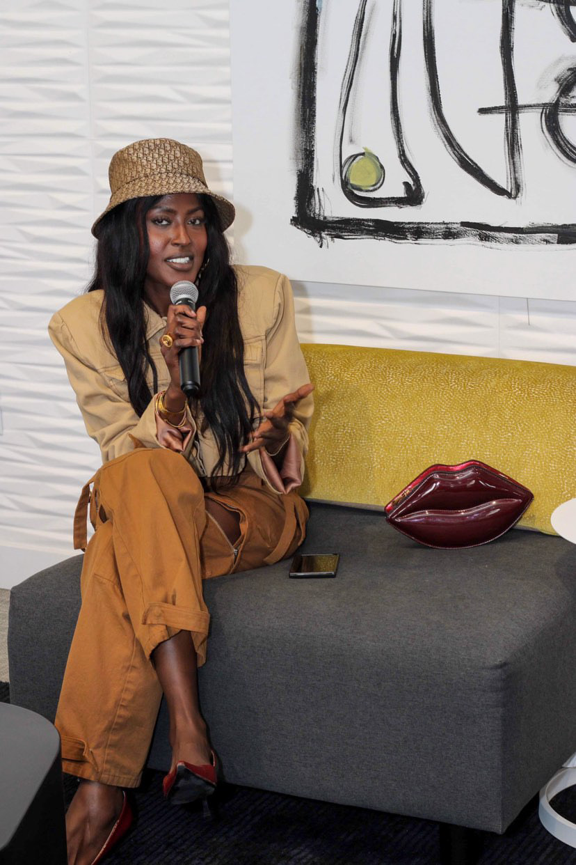 Woman with long dark hair, wearing a hat and beige jacket, sitting on a yellow and gray sofa, holding a microphone.
