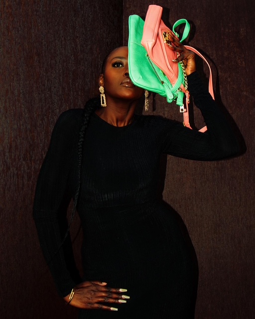 Woman with dark skin holding colorful handbag above her head, wearing a black outfit, against dark background.