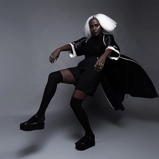 Person with white hair poses in black clothing against a gray background, one leg extended, arms outstretched, dramatic lighting.
