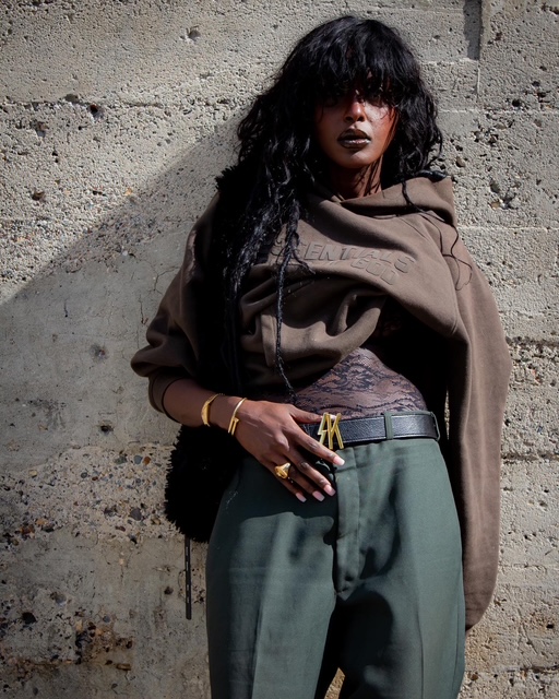 Woman with dark curly hair and dark lipstick stands against a textured wall, wearing a brown hoodie and green pants.