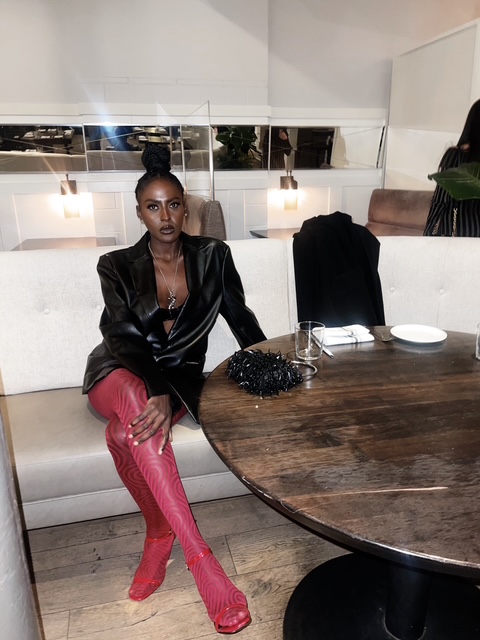 Woman sitting at a table in a restaurant, wearing a black jacket and red pants, with a purse and a plate on the table.