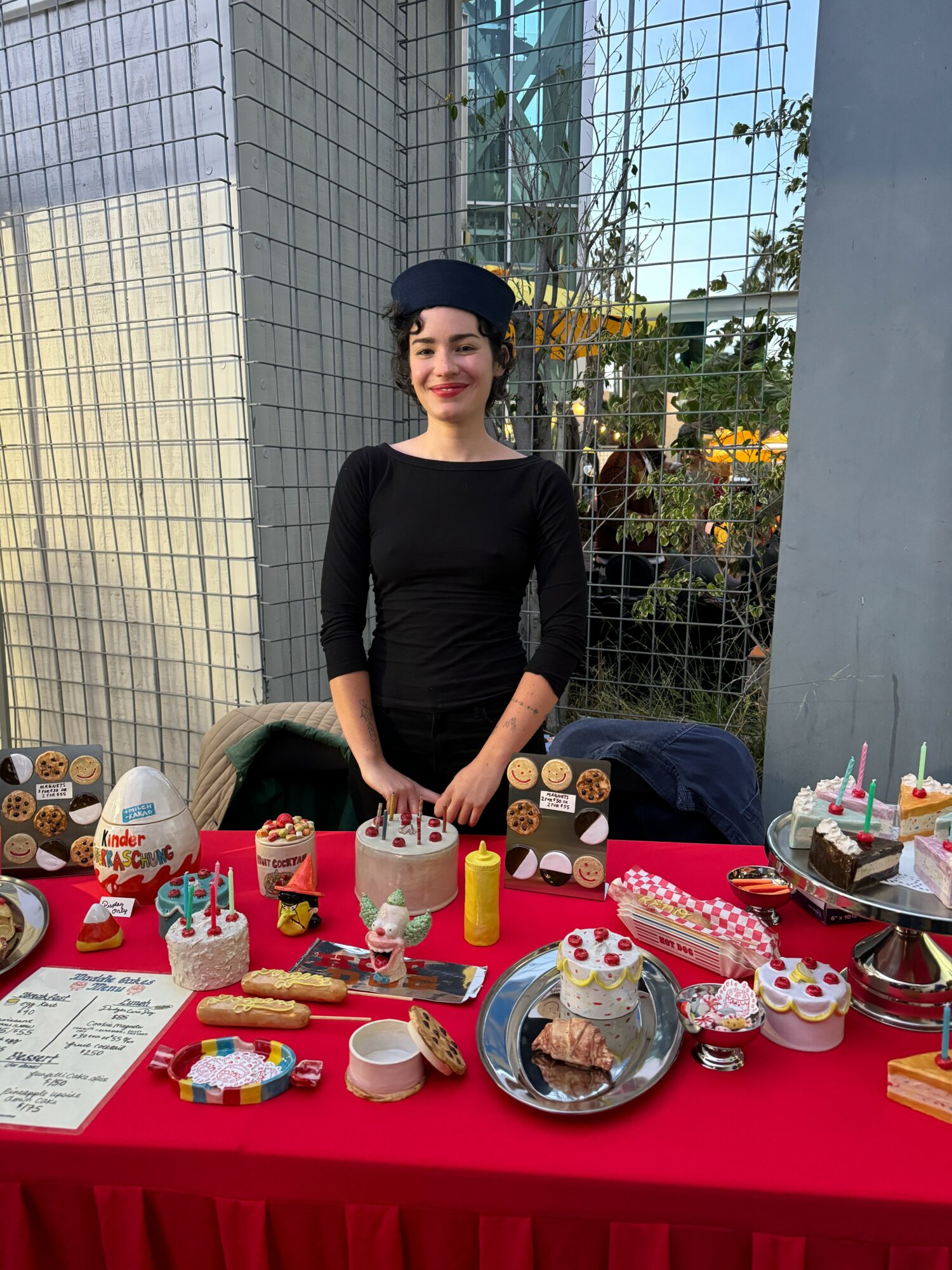 Woman standing behind a table with birthday cakes and treats outdoors, smiling, wearing a black dress and hat.
