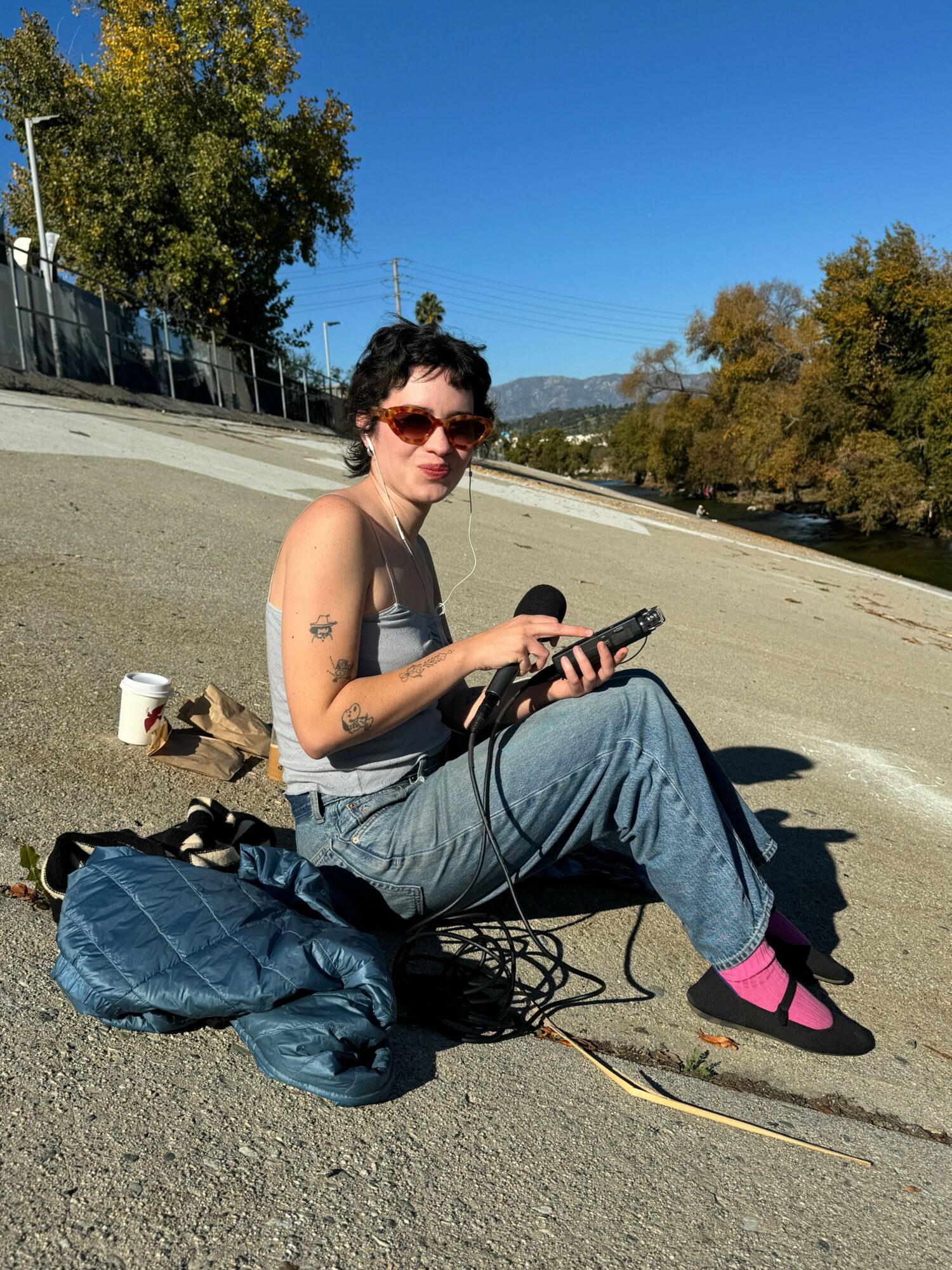 Person sitting on the ground with sunglasses, holding a phone, with a bag and shoes nearby, outdoors on a sunny day.