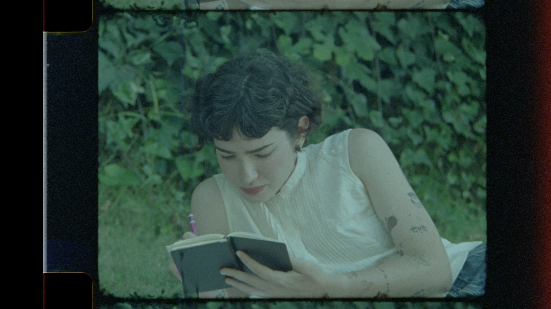 Young woman with short dark hair reading a book outdoors, surrounded by green foliage.