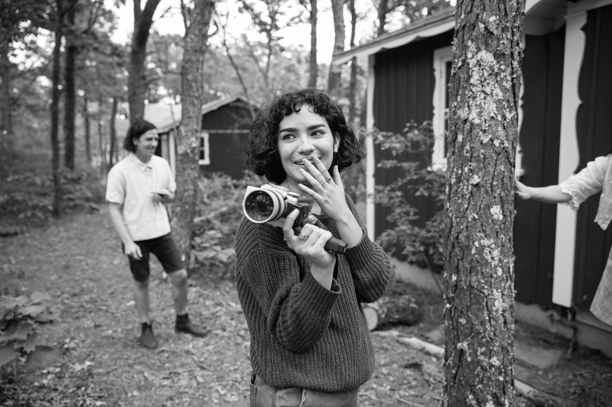 Young woman with curly hair holding a camera, smiling, in a wooded outdoor area with two other people nearby.