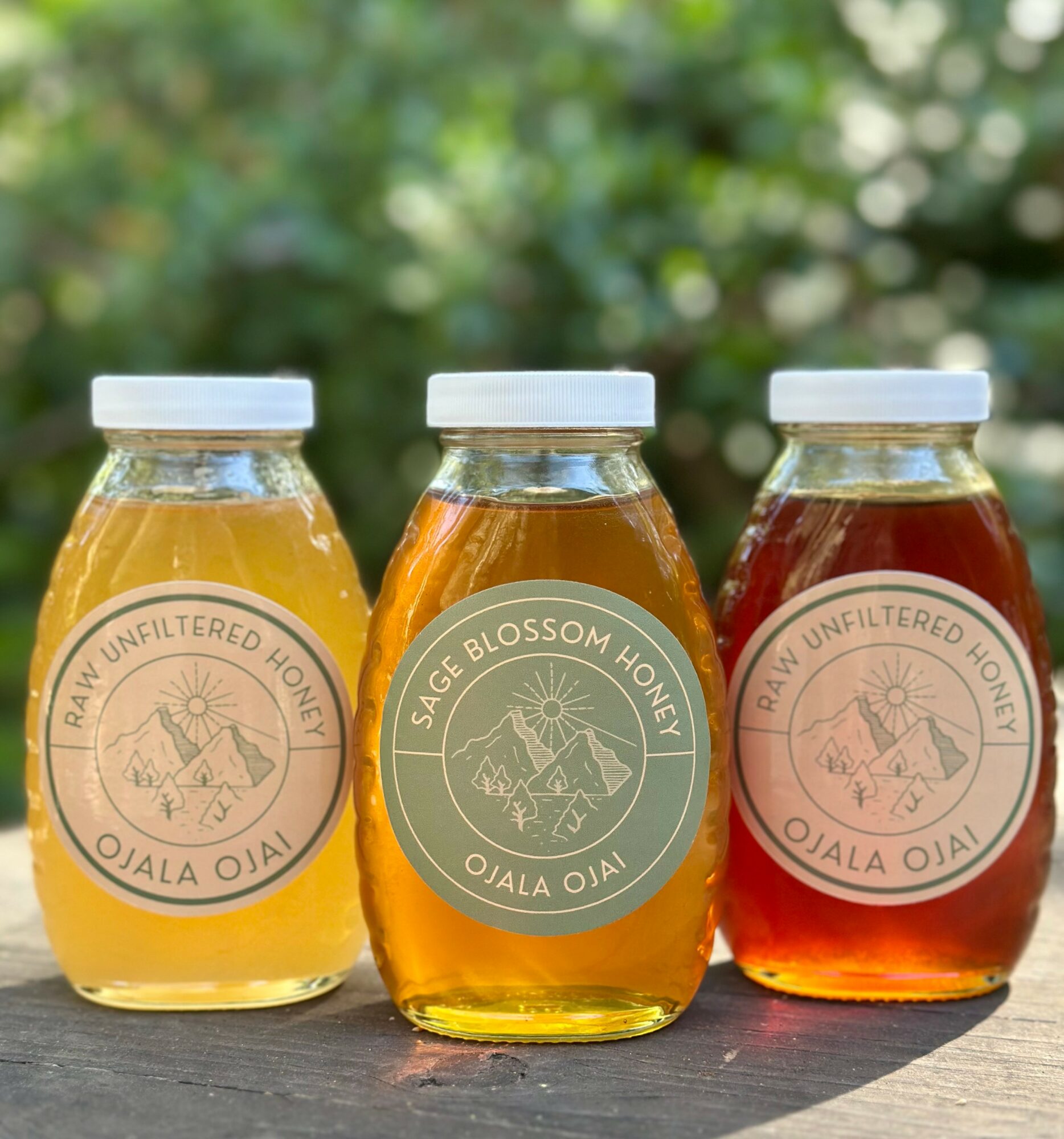 Three jars of honey with white lids, labeled with different honey types, placed outdoors on a wooden surface, with a blurred green background.