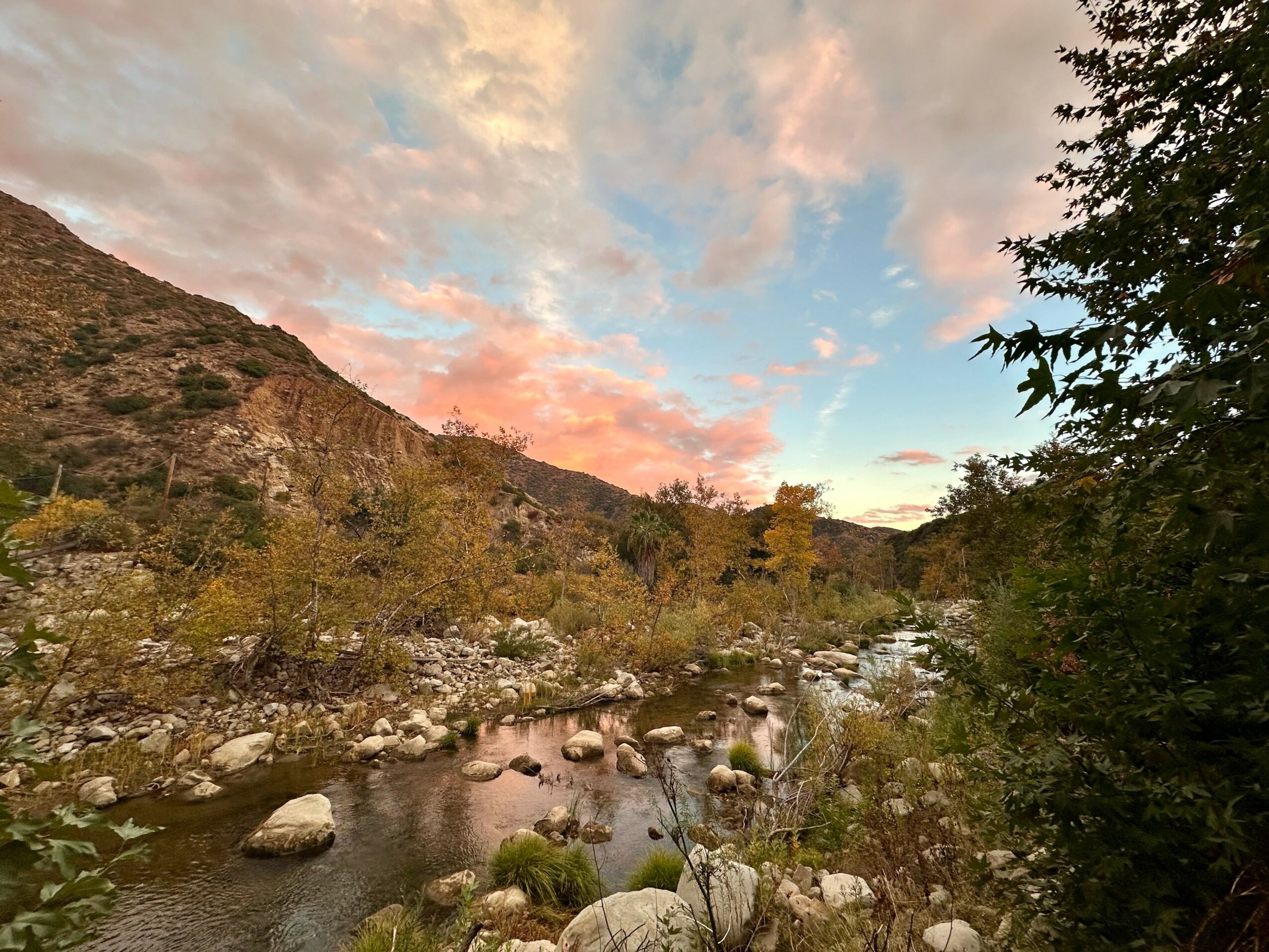 River flowing through a rocky landscape with trees and a mountain under a colorful sky at sunset.