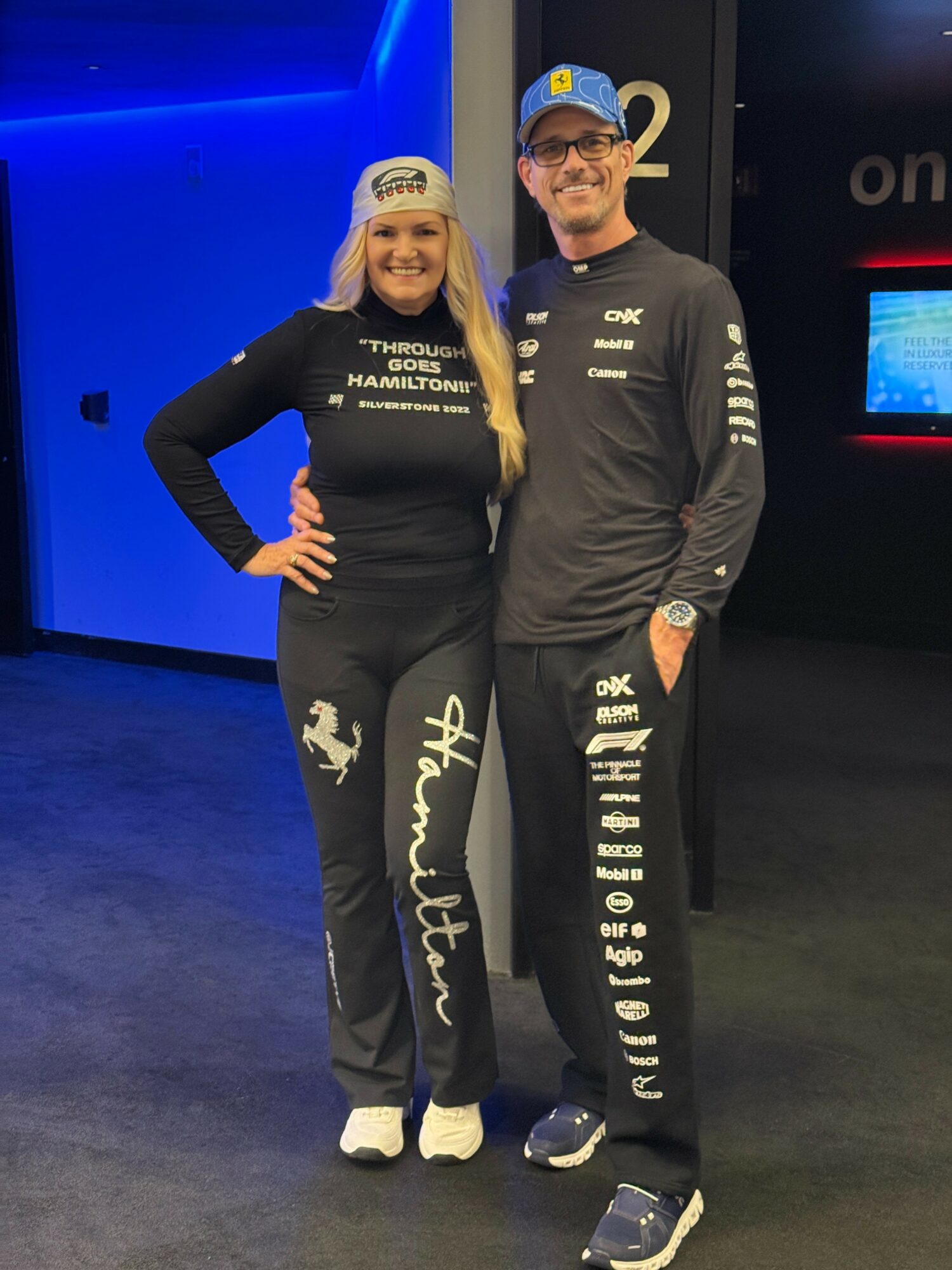 Two people stand together in a dark room with blue lighting, smiling at the camera. The woman wears a black outfit with white text and a cap, and the man wears a black outfit with white text and glasses.