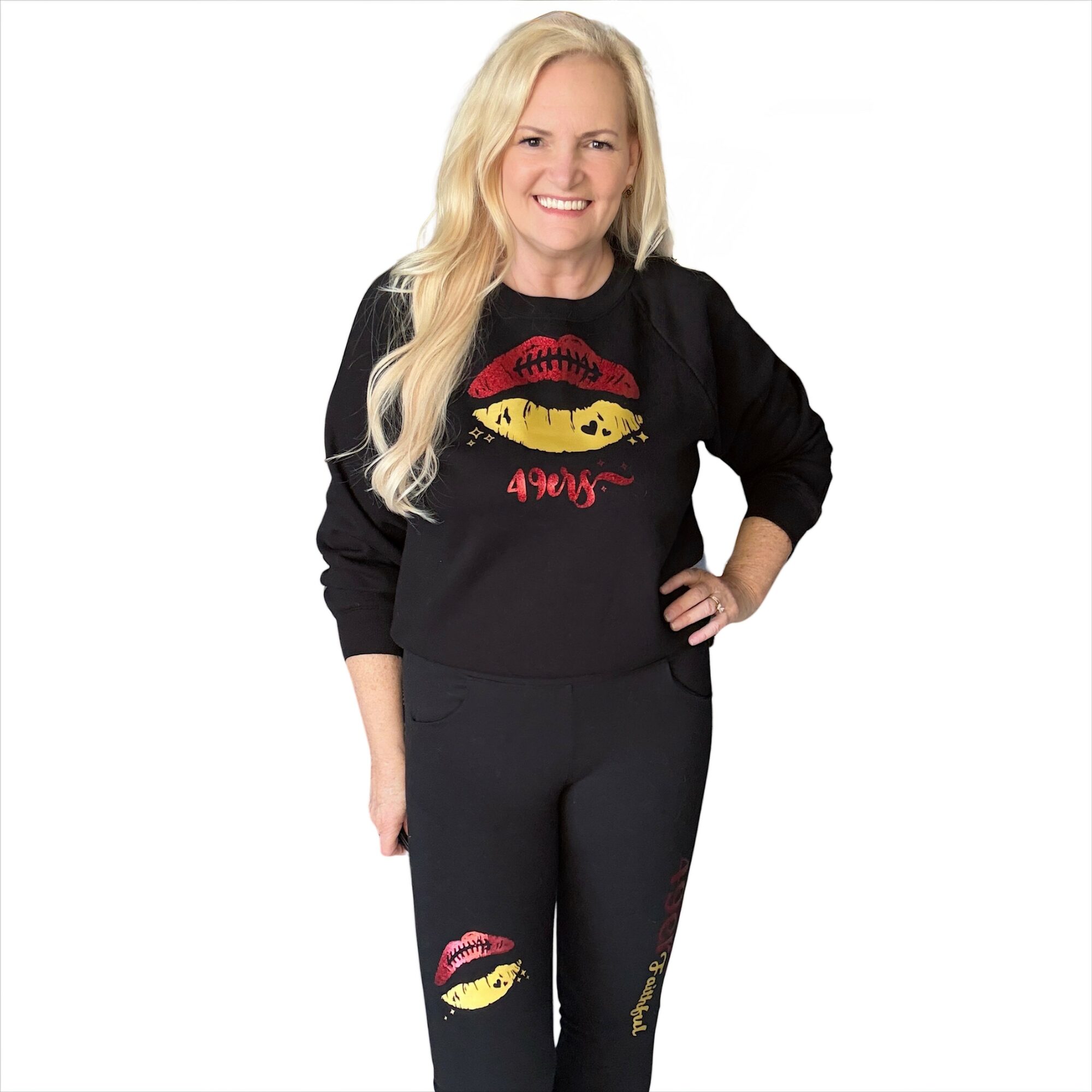 Woman with long blonde hair smiling, wearing a black sweatshirt and pants with yellow lips and red text, standing with hand on hip.